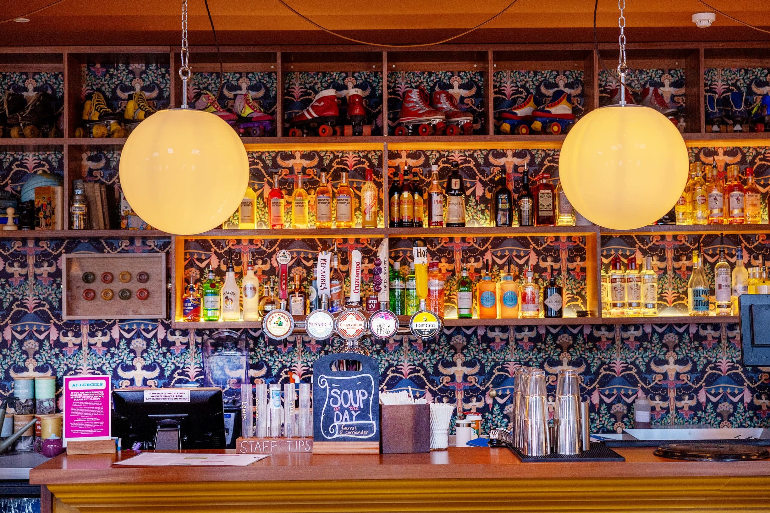 A colorful durano bar with two glowing round pendant lights, bottles of liquor on shelves, several beer taps, a cash register, and a "Soup of the Day" sign displayed on the counter. A vibrant, patterned wall is in the background.
