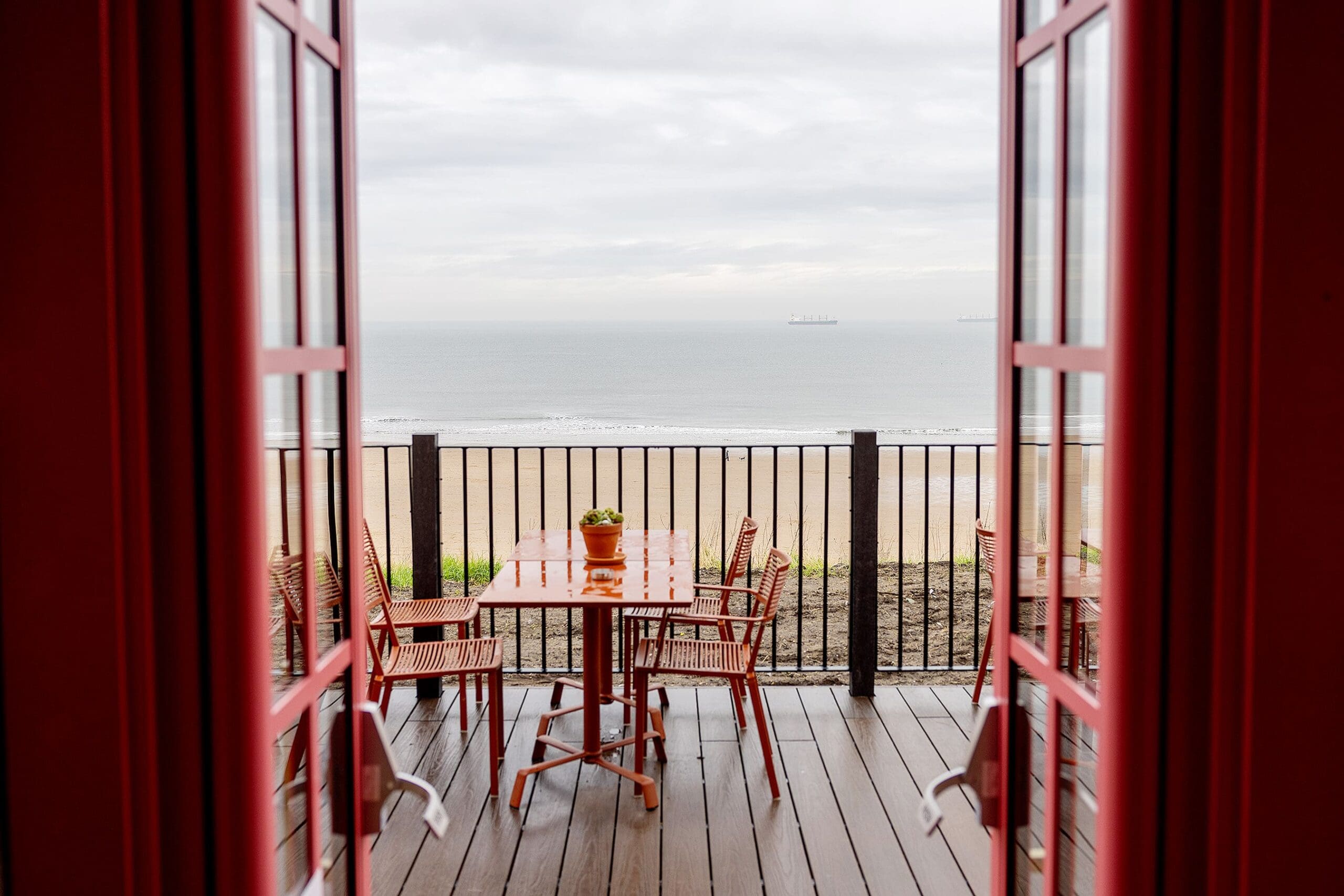 A view through open red-framed doors to a balcony with a wooden table and chairs overlooking a sandy durano beach and calm sea under a cloudy sky.