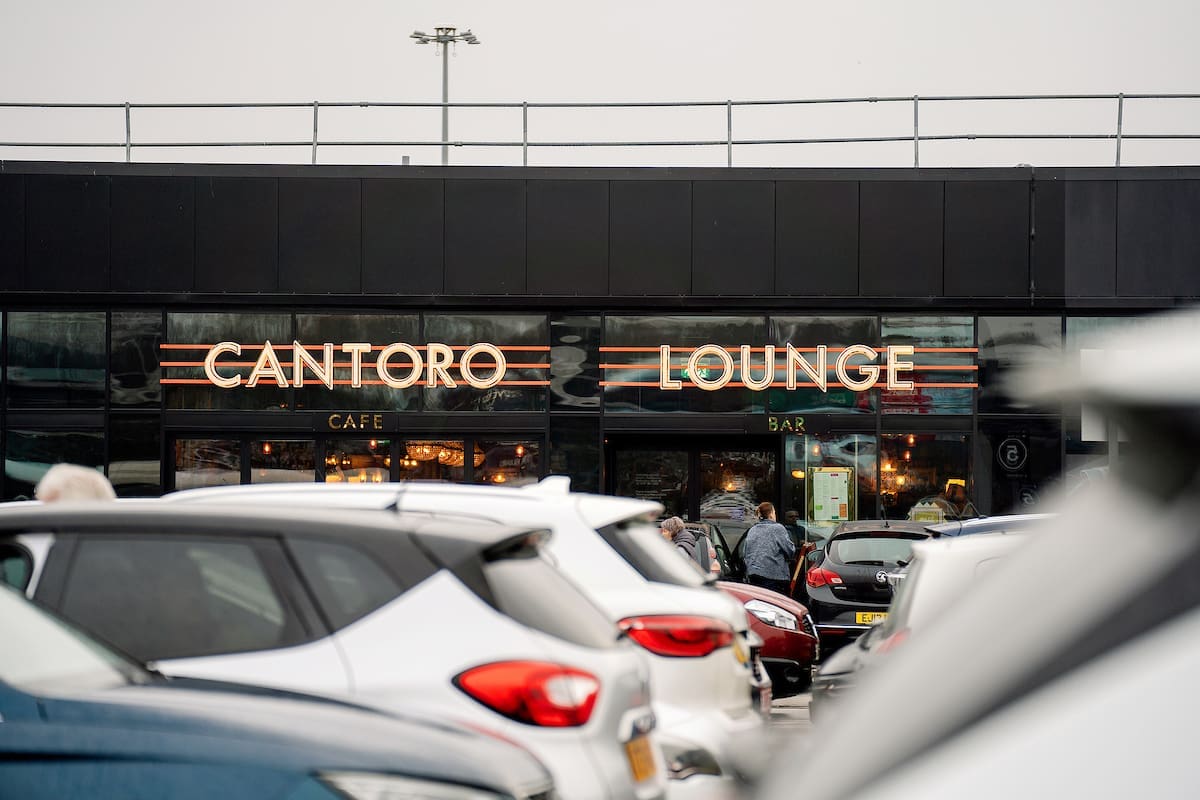 A row of parked cars lines the street in front of Cantoro Lounge, a modern building with large windows and glowing neon signs. The softly lit interior reveals people enjoying their evening inside.