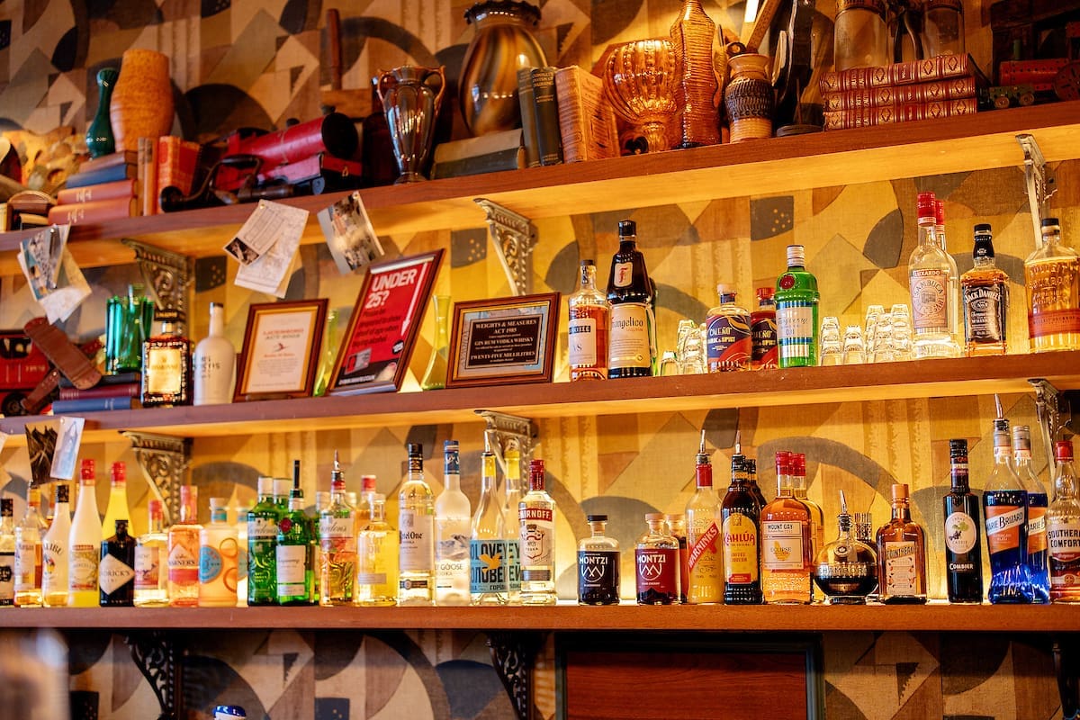 A bar shelf lined with various bottles of liquor, spirits, and Cantoro mixers, with awards, glasses, and vintage decor displayed against a patterned wallpaper background.
