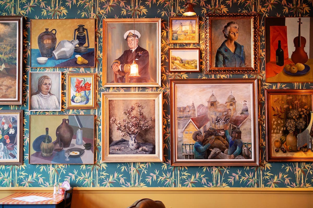 A wall covered with framed paintings—including portraits, still lifes, buildings, and musical instruments—creates a cozy Cantoro vibe against green wallpaper with a yellow leaf pattern. Warm lighting adds to the inviting atmosphere.