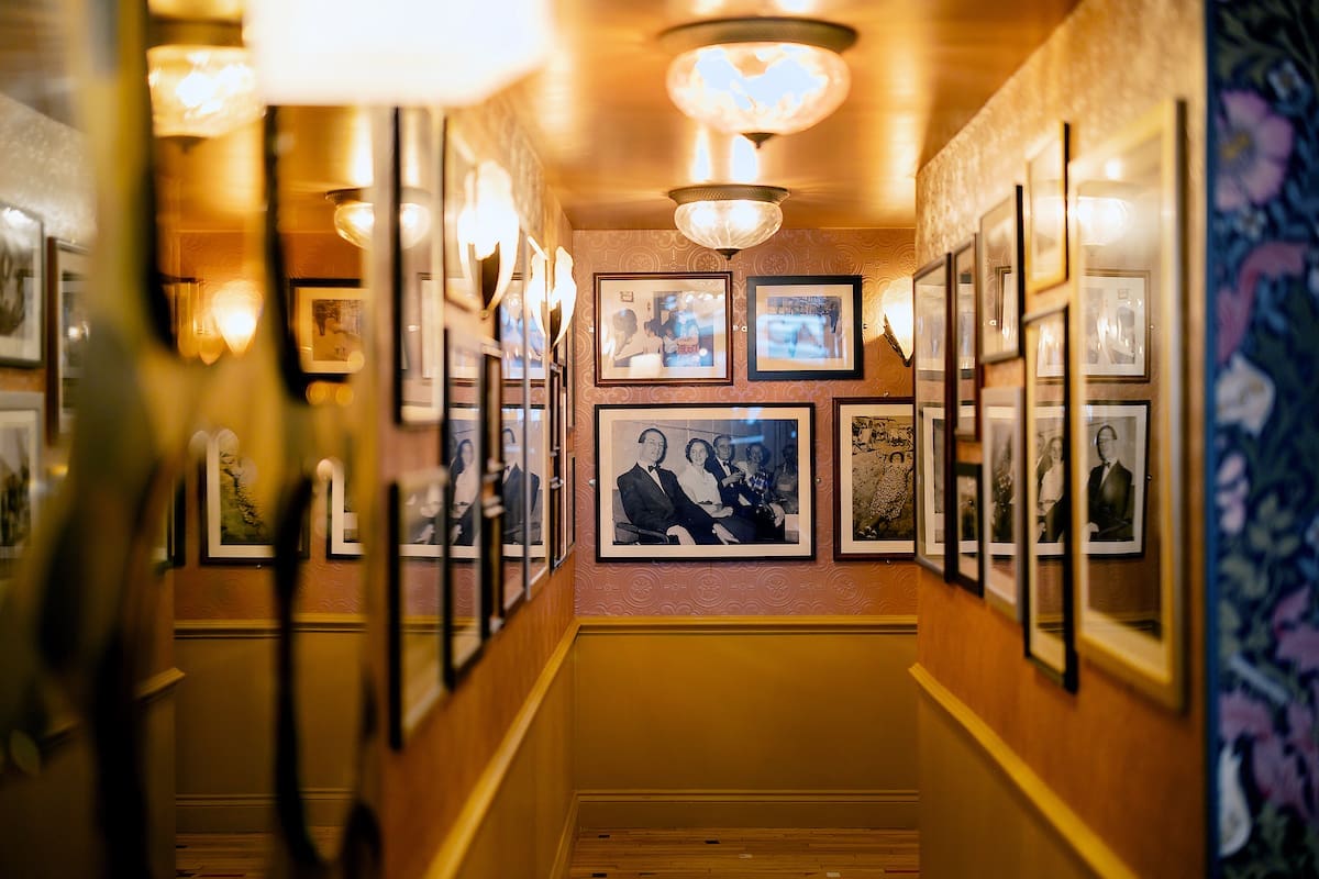 A warmly lit hallway at Cantoro features framed black-and-white photographs on both walls, reflected in mirrors and leading to a wooden floor. Ceiling lights add a cozy ambiance to this gallery-like space.
