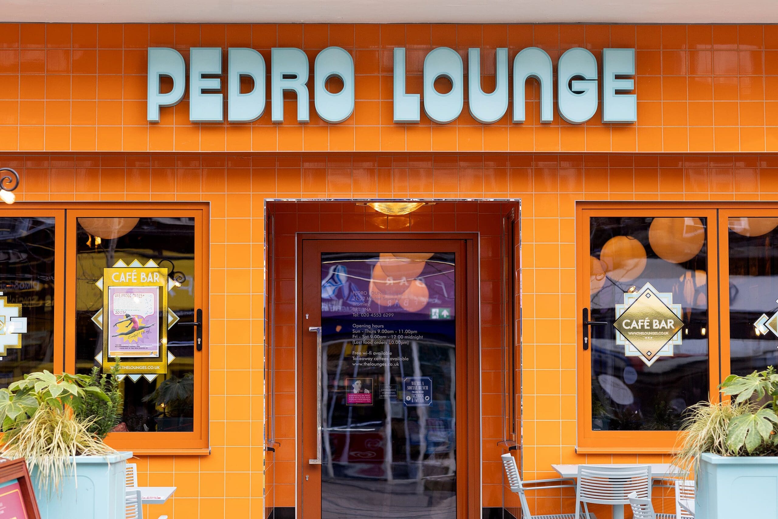 A storefront with bright orange tiles, blue-framed windows, and a sign reading “Pedro Lounge” above the entrance. Posters fill the windows, while outdoor tables and potted plants create a welcoming vibe at Pedro’s spot.