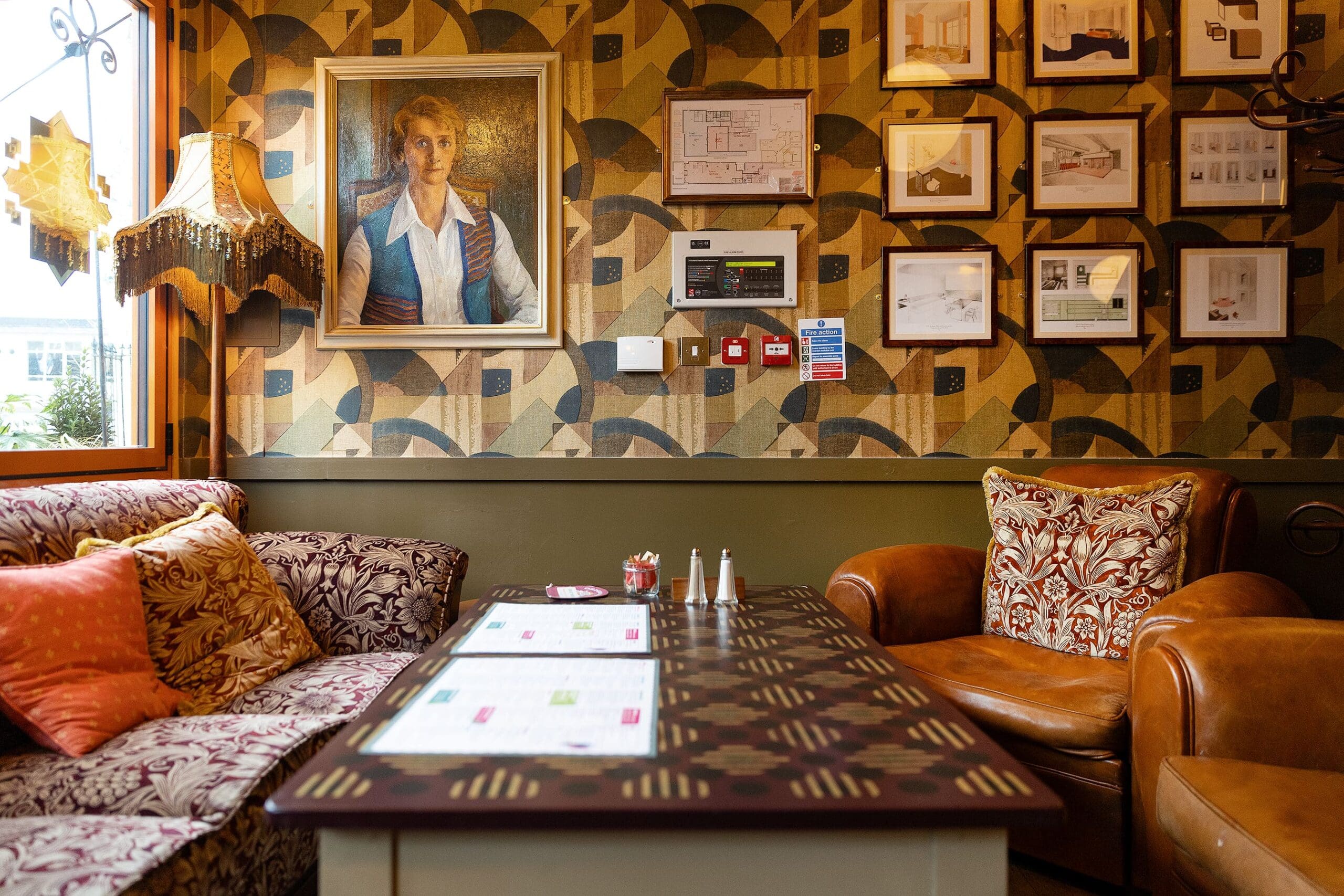 Cozy vintage lounge with patterned sofas and armchair, a geometric wallpaper, framed art and a portrait of Pedro, a table with menus, and a decorative lamp by the window. The ambiance is warm and inviting.