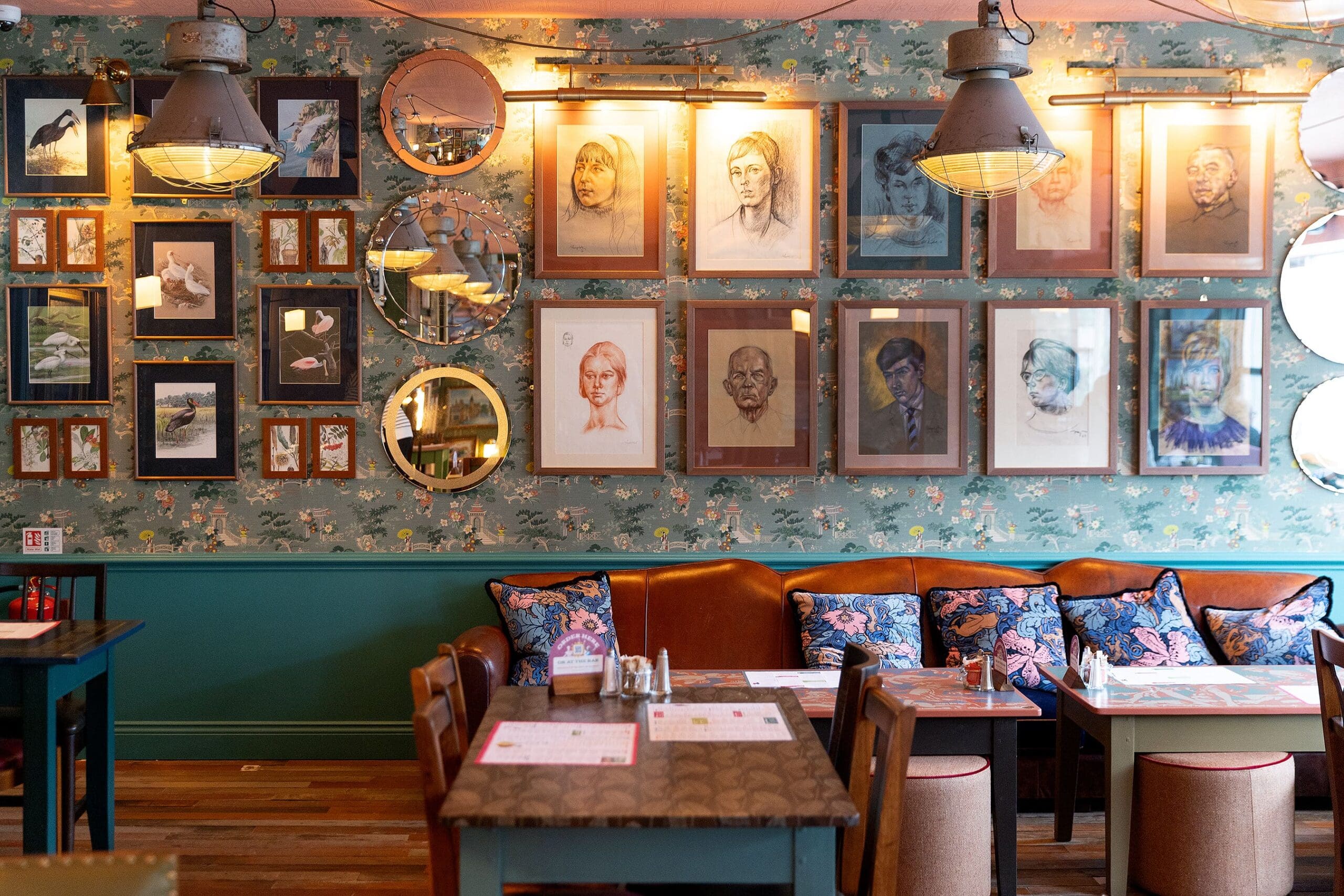 A cozy restaurant with wooden tables and brown leather booths, decorated with throw pillows. The wall displays framed portraits, bird illustrations by Pedro, and round mirrors, with floral-patterned wallpaper and industrial-style lights above.