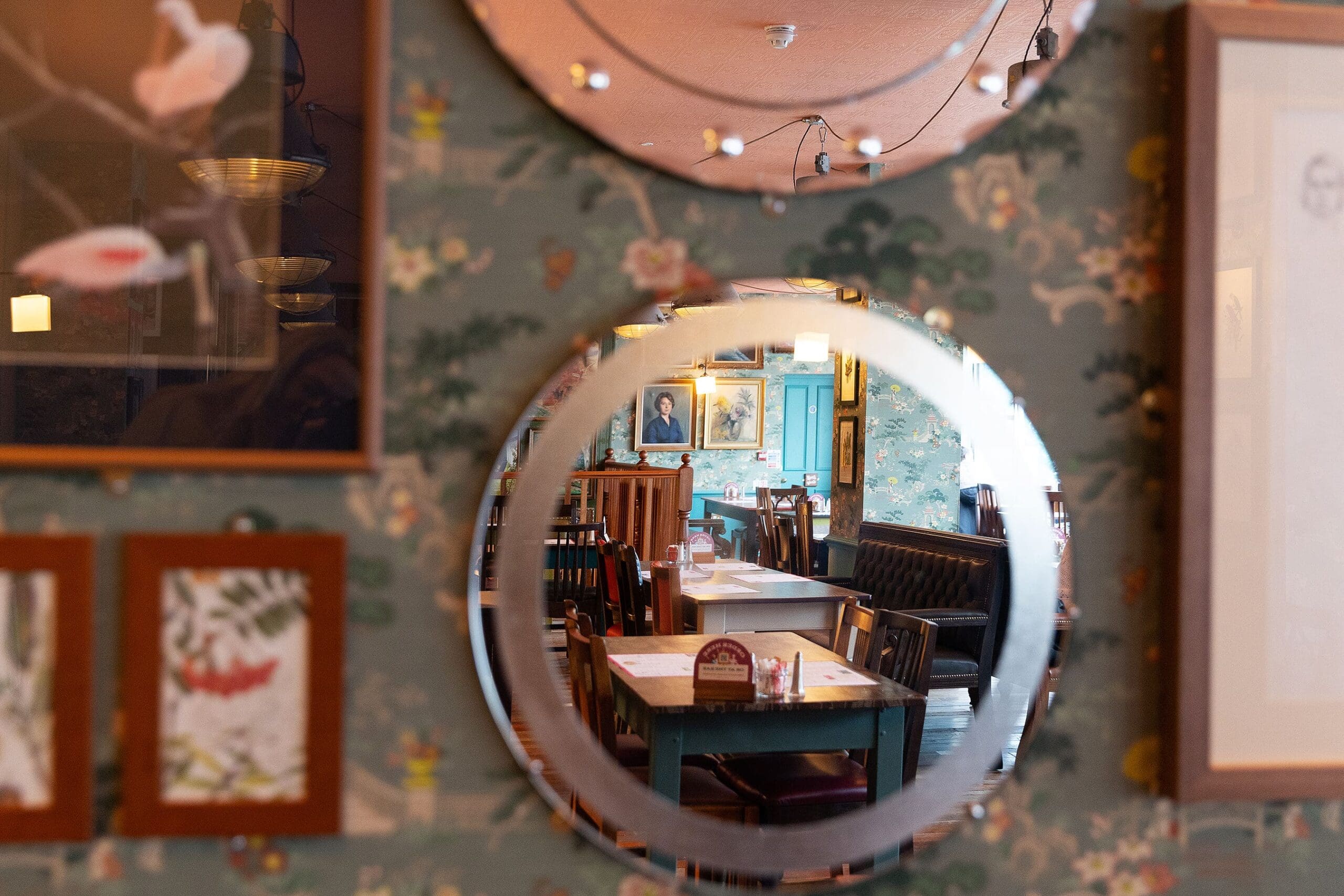 A round mirror on a floral-patterned wall reflects Pedro’s cozy restaurant interior with wooden tables, chairs, booths, and framed pictures, creating a warm, inviting atmosphere.