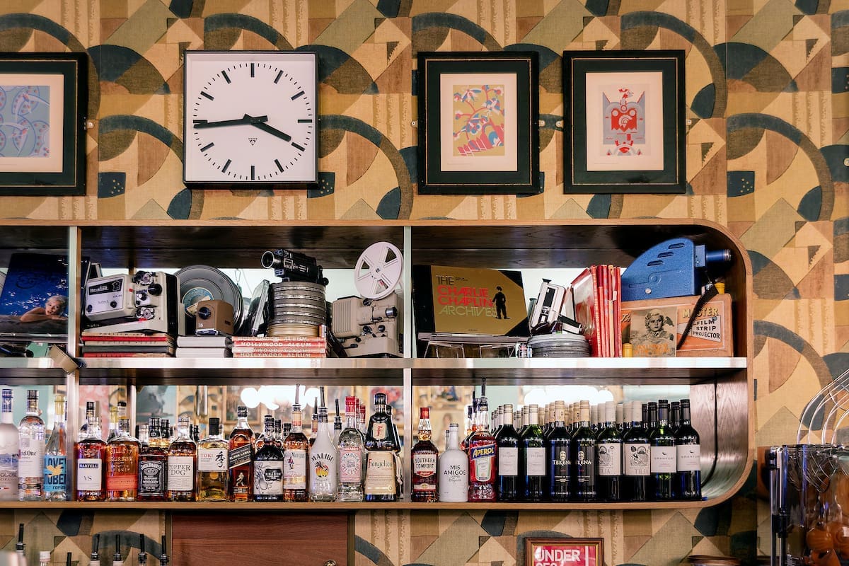 A bar shelf displays various liquor bottles, wine, calvero memorabilia, vintage film equipment, books, and keepsakes. Above the shelf is a large clock and framed art, all set against a retro-patterned wallpaper.