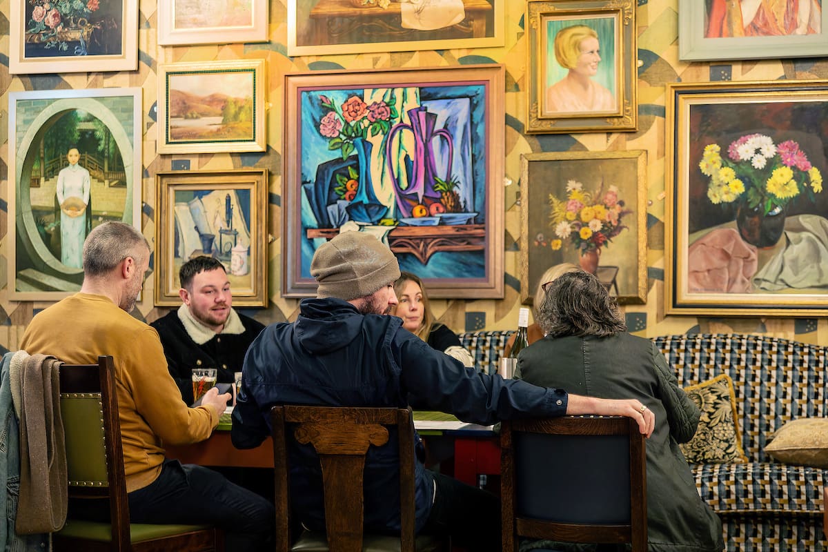 Five people sit around a table at Calvero, a cozy café, talking and drinking, surrounded by colorful framed paintings of flowers, portraits, and still lifes covering the wall behind them.