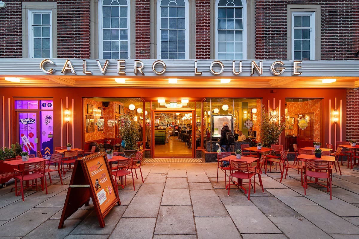 Outdoor view of Calvero Lounge café with red chairs and tables set on a stone patio, bright lighting inside, large windows, and a welcoming calvero atmosphere, with one person standing near the entrance.