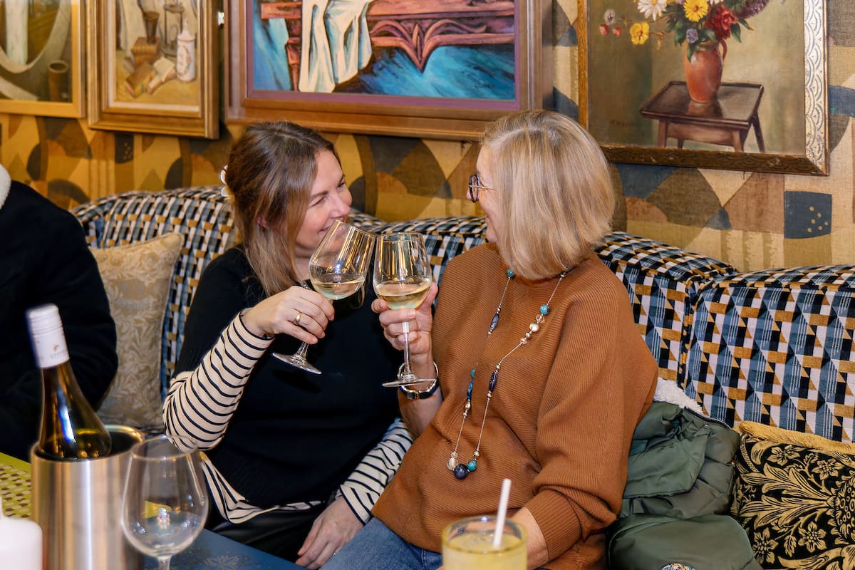 Two women sit on a patterned couch, smiling and clinking glasses of calvero white wine. The background features colorful framed paintings and patterned wallpaper. One woman wears a brown sweater, the other a striped shirt.
