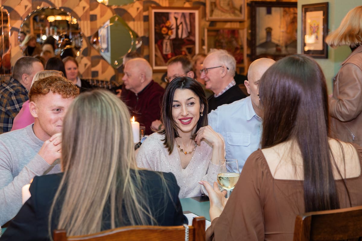A group of people sit at a restaurant table, talking and smiling. The lively room is filled with diners and Calvero’s colorful, artistic decor. Framed art lines the walls as one woman holds a glass of white wine.