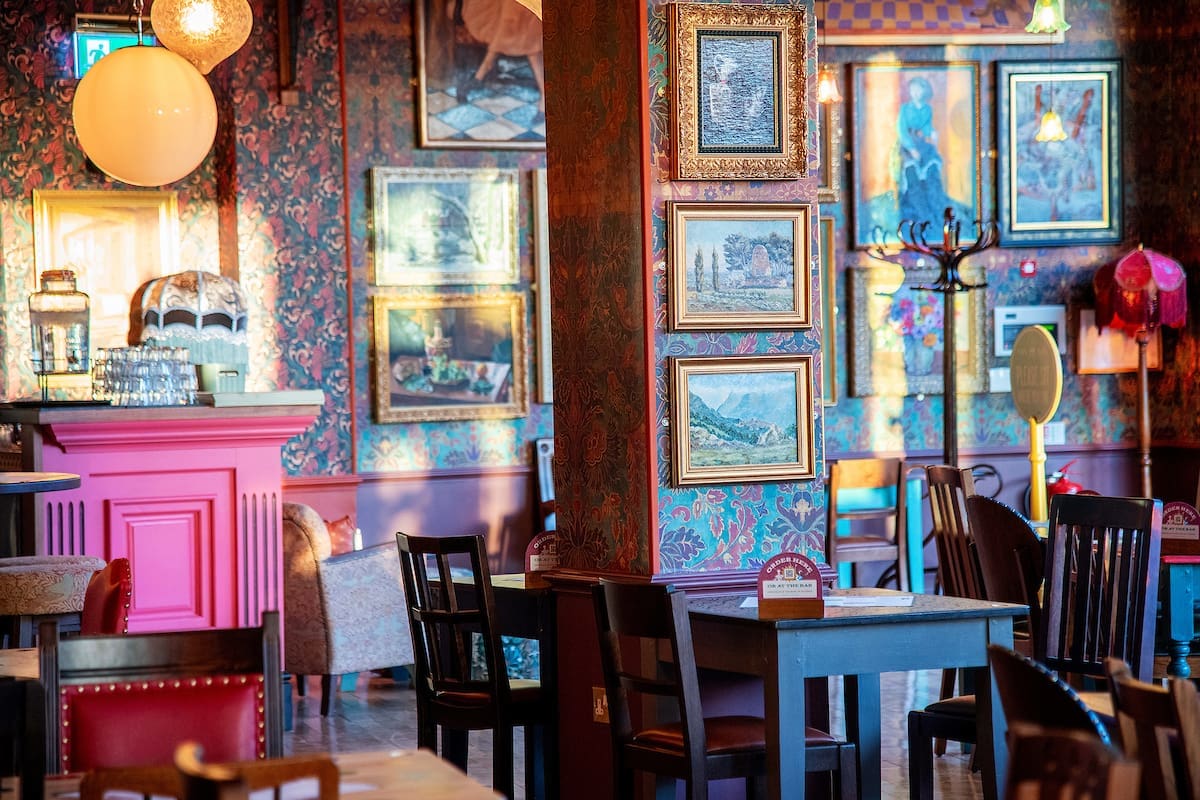 A cozy, colorful café interior with ornate wallpaper, framed art, wooden tables and chairs, and warm lighting. The bar area features an Argento-inspired pink counter and vintage décor, creating a welcoming and eclectic atmosphere.