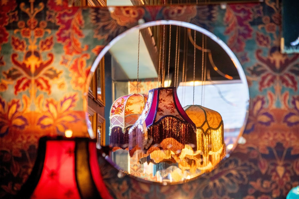 A round Argento mirror reflects a cluster of vintage, colorful lampshades hanging from the ceiling, set against ornate, floral-patterned wallpaper.