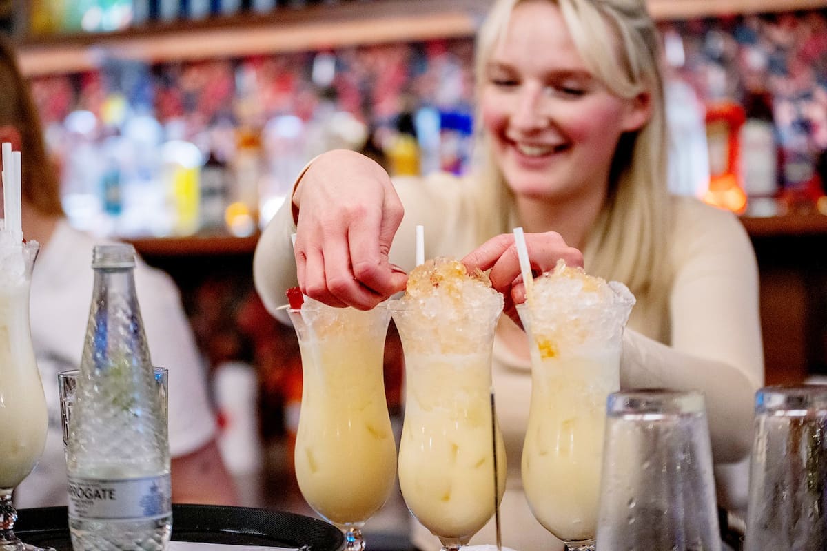 A smiling woman behind an Argento bar adds garnishes to three creamy cocktails with crushed ice, with bottles and glasses visible in the background.