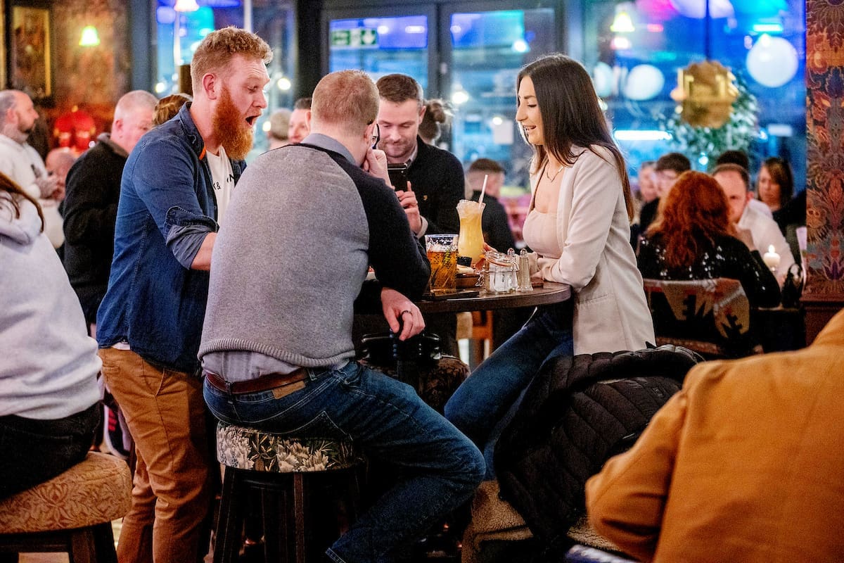 A group of four people sit and stand around a high table in Argento, a busy, colorful bar, chatting and smiling with drinks in front of them. Other patrons are visible in the background.