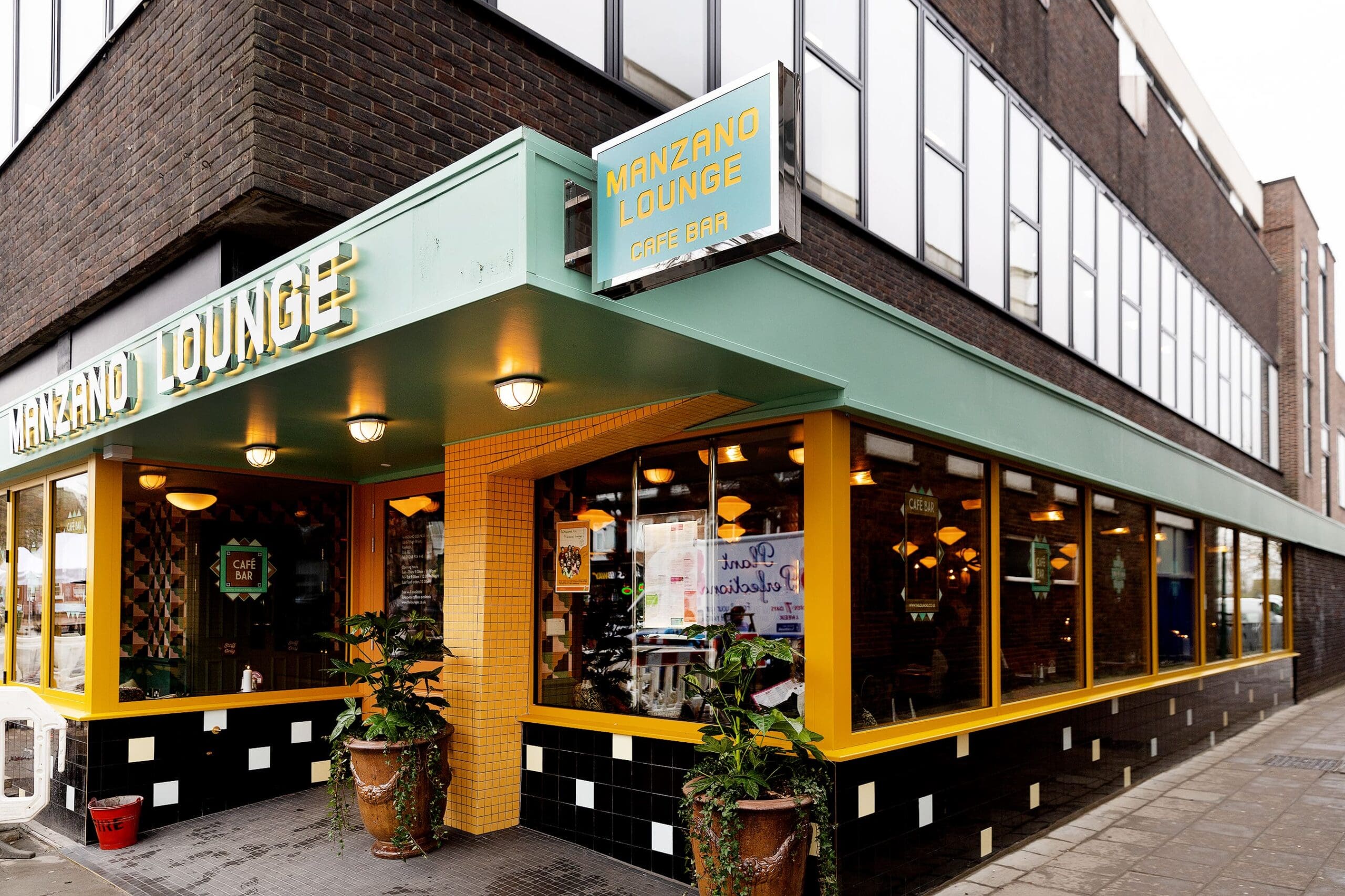 The exterior of Manzano Lounge café bar, featuring large windows, manzano-yellow trim, green signage, and potted plants by the entrance on a city sidewalk.