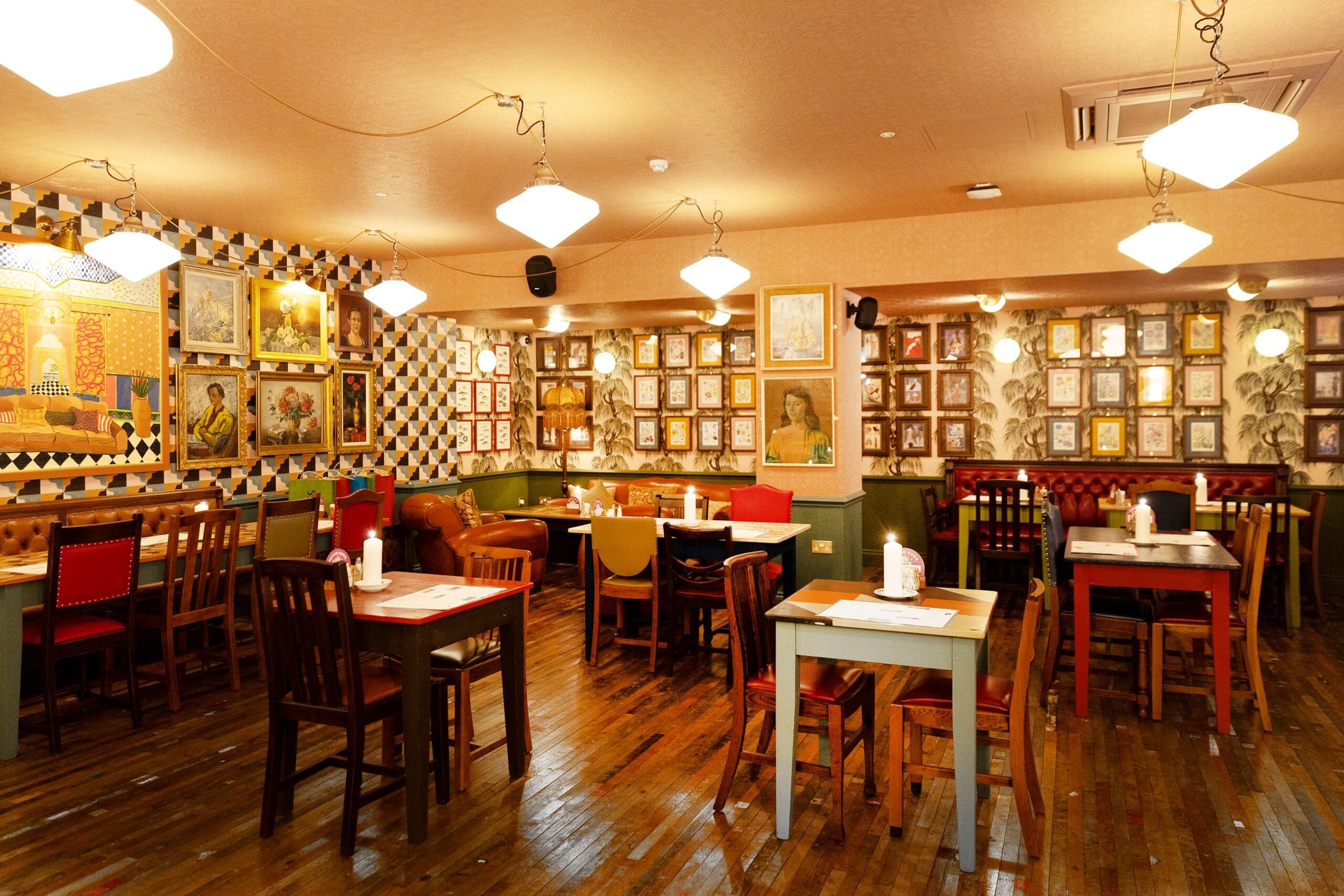 A cozy, warmly lit restaurant interior with wooden floors, colorful mismatched chairs, and walls adorned with framed art and photos—perfect for enjoying a meal at Manzano. Several tables are set and ready, but no people are present.