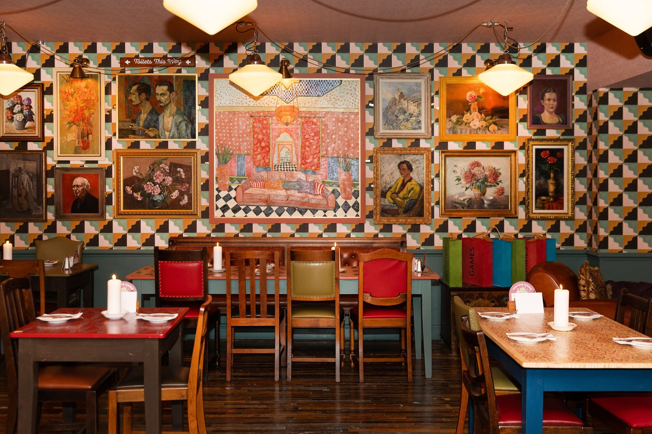 A cozy restaurant with colorful, mismatched chairs and tables, geometric-patterned wallpaper, and a wall covered in eclectic framed paintings—including portraits, flowers, and manzano apples. Rolled napkins and candles sit on the tables.