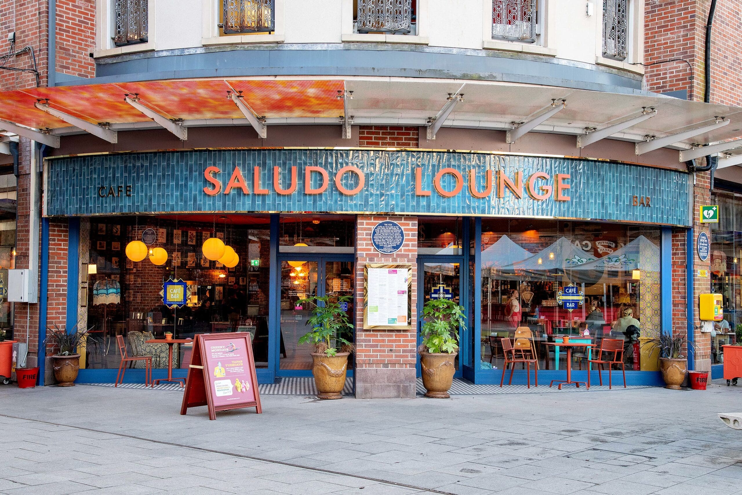 Street view of Saludo Lounge café and bar, with a blue tiled saludo facade, large windows showing a cozy interior with tables and hanging lights, plus outdoor seating featuring red and teal chairs.