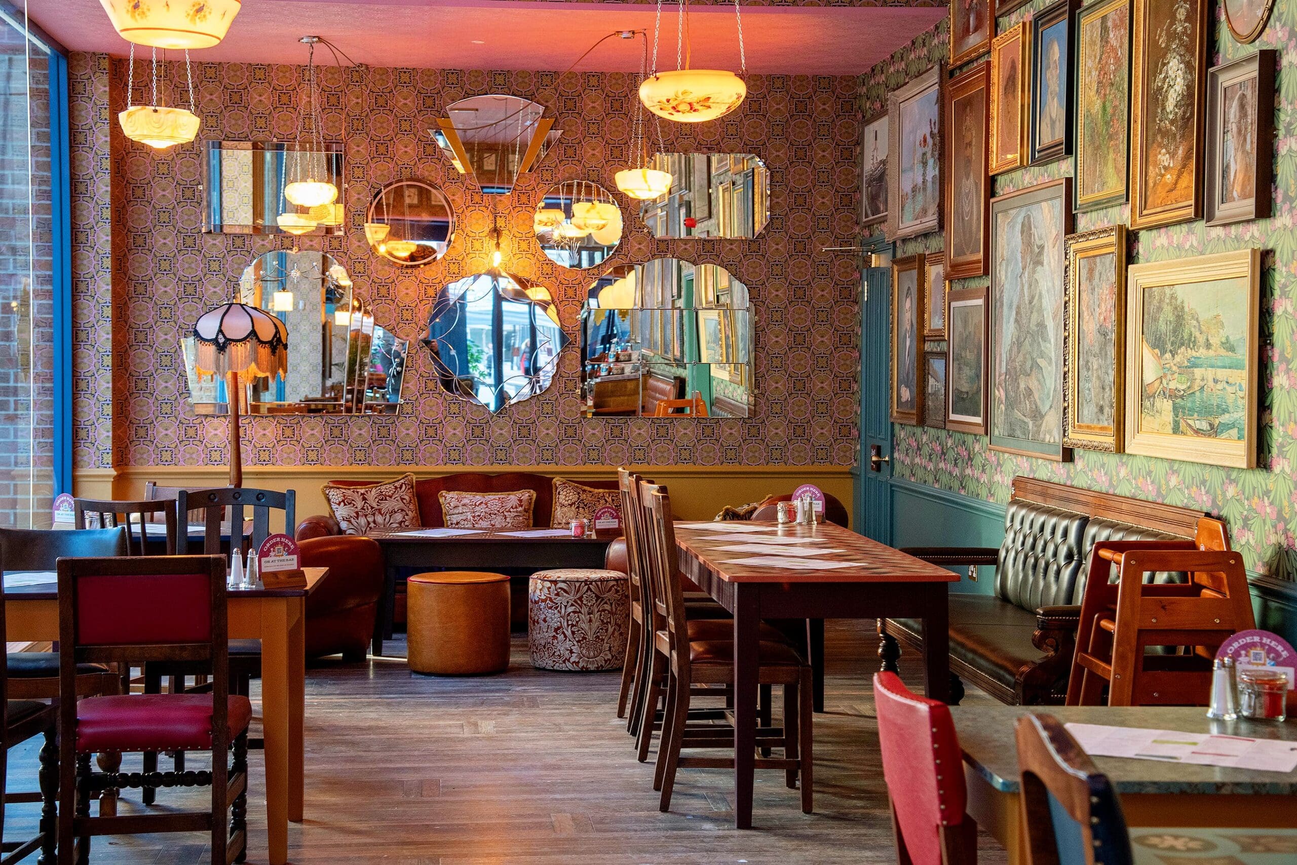 Eclectic café interior with mismatched tables, vintage chairs, framed artwork on patterned walls, and an arrangement of decorative mirrors reflecting warm pendant lights. A retro vibe and cozy saludo welcome guests into this inviting space.