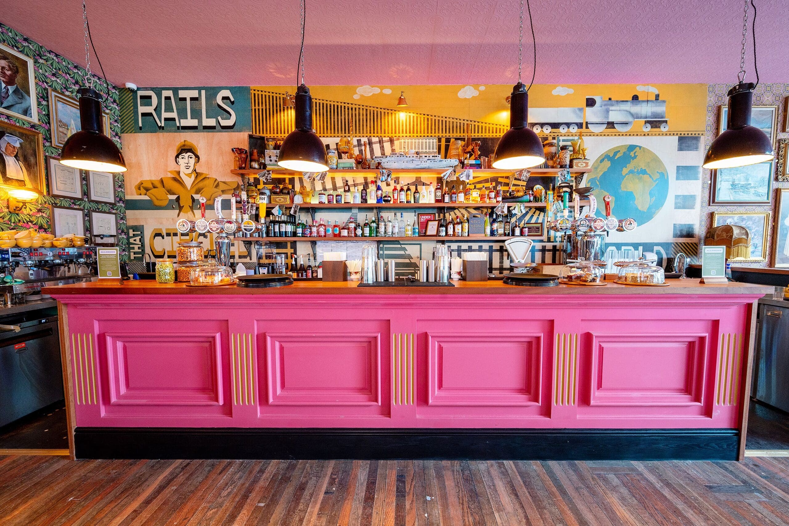 A vibrant, eclectic bar with a bright pink counter, hanging black lamps, and colorful wall art—including a globe, a "RAILS" sign, and a cheerful saludo to guests. Shelves behind the bar are stocked with bottles and glassware.