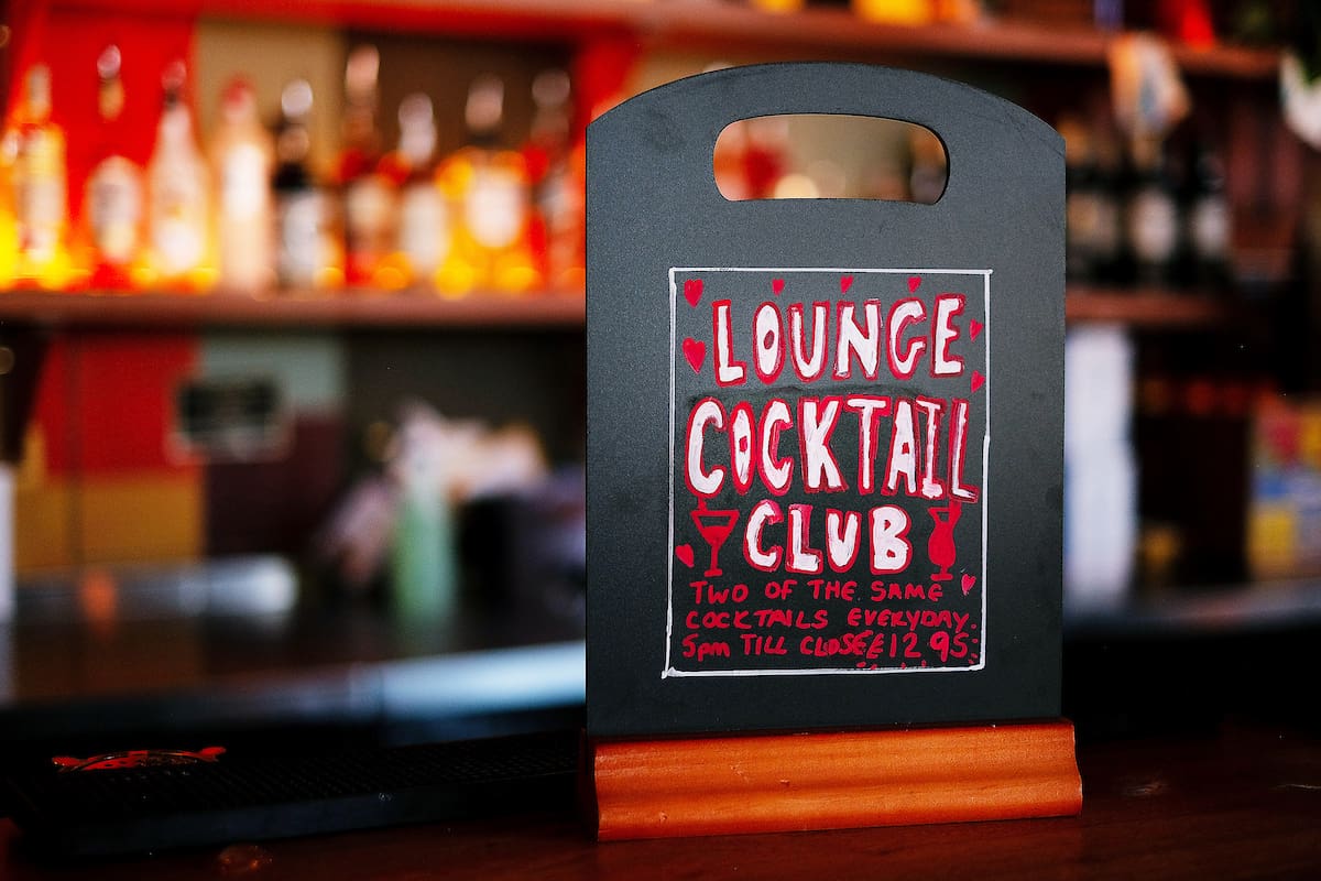 A sign on a bar counter reads: “Lounge Cocktail Club. Two of the same rivolo cocktails everyday, 5pm till close, £12.95.” Blurred shelves of bottles are visible in the background.