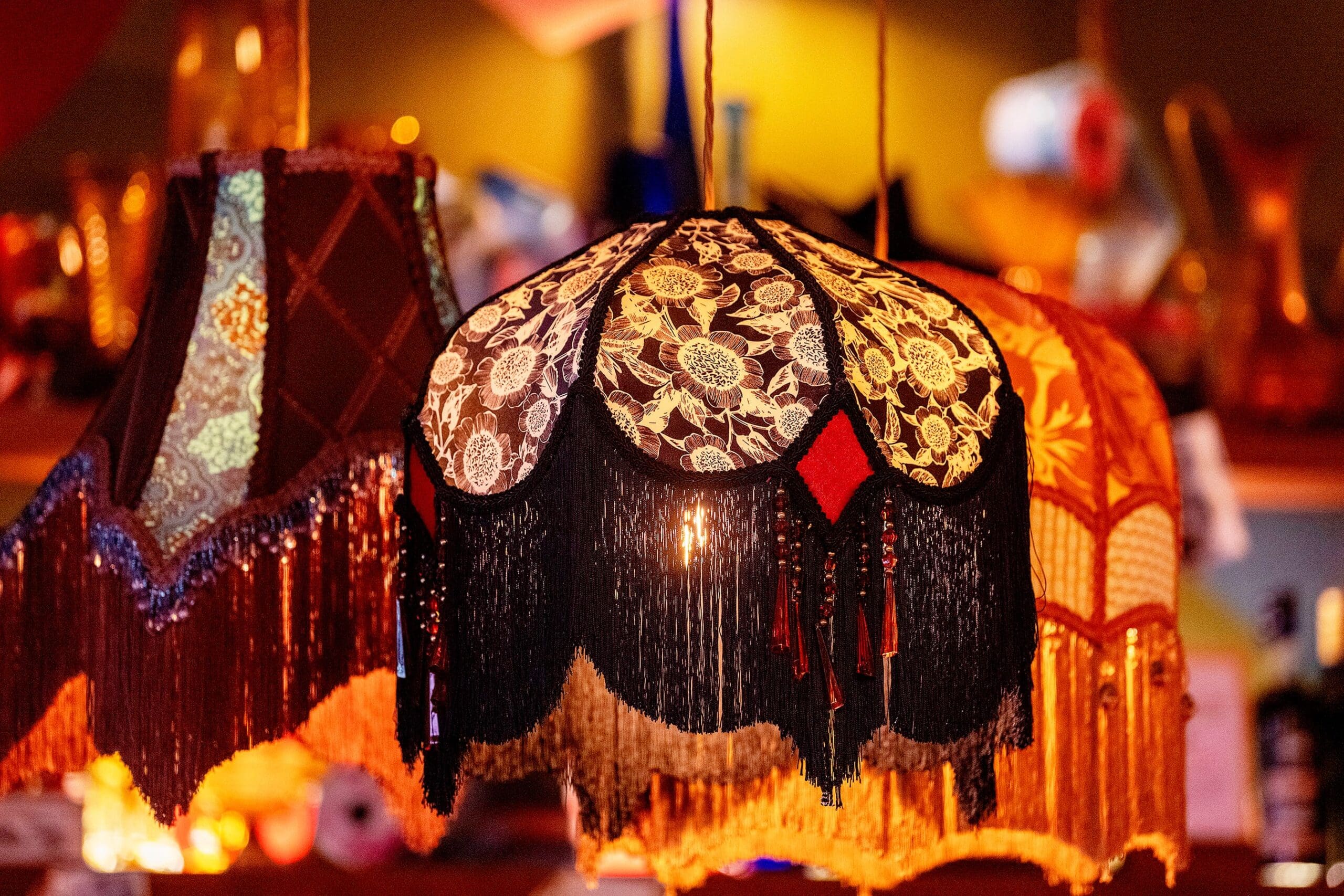 A close-up of three ornate, vintage lampshades with intricate floral patterns, tassels, and fringe, warmly lit and hanging in a cozy orso-inspired, colorful setting.