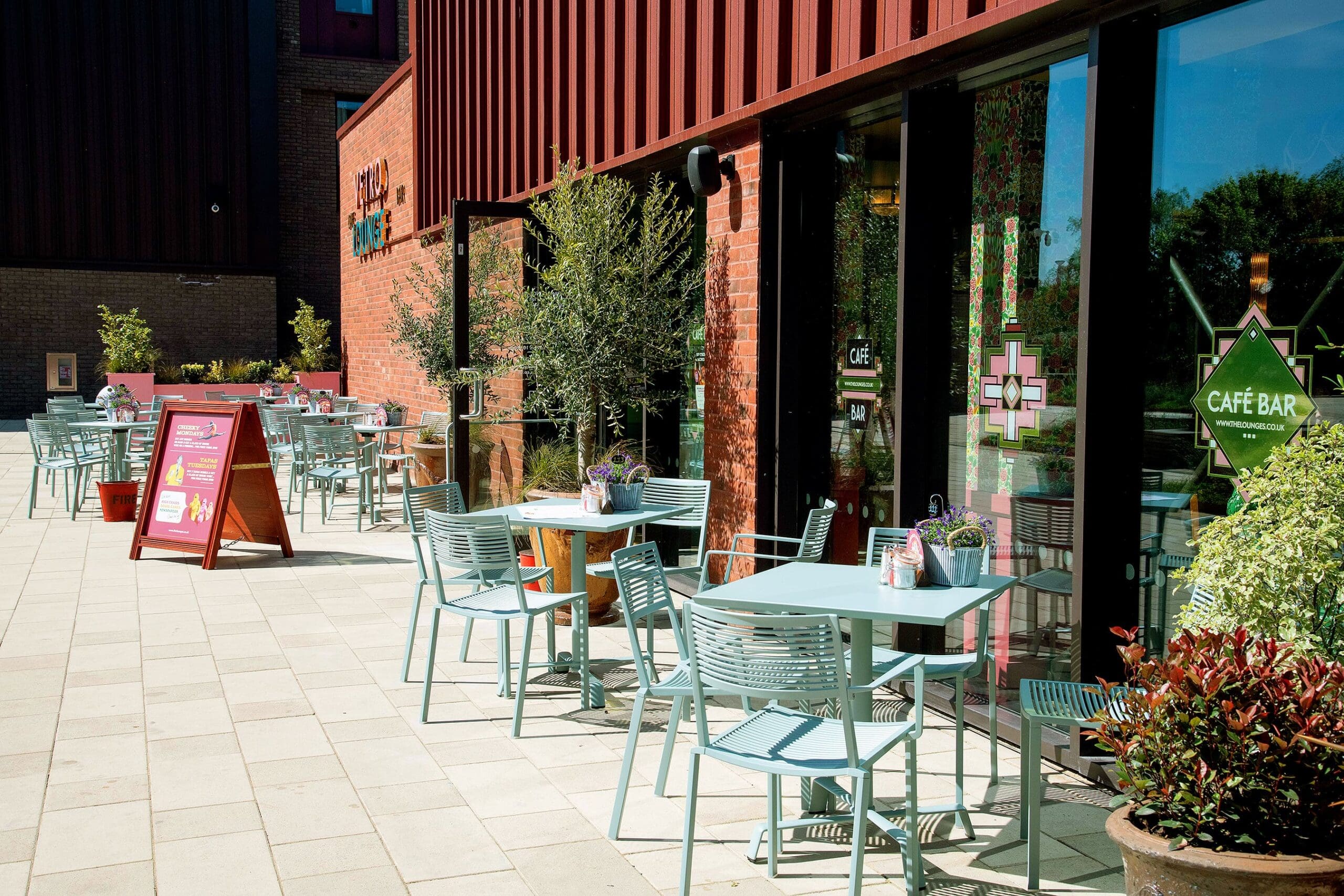Outdoor seating area of Vetro café with light blue chairs and tables, potted plants, and a menu board on a sunny day. Large windows reflect greenery, and the Vetro café sign is visible on the brick wall.