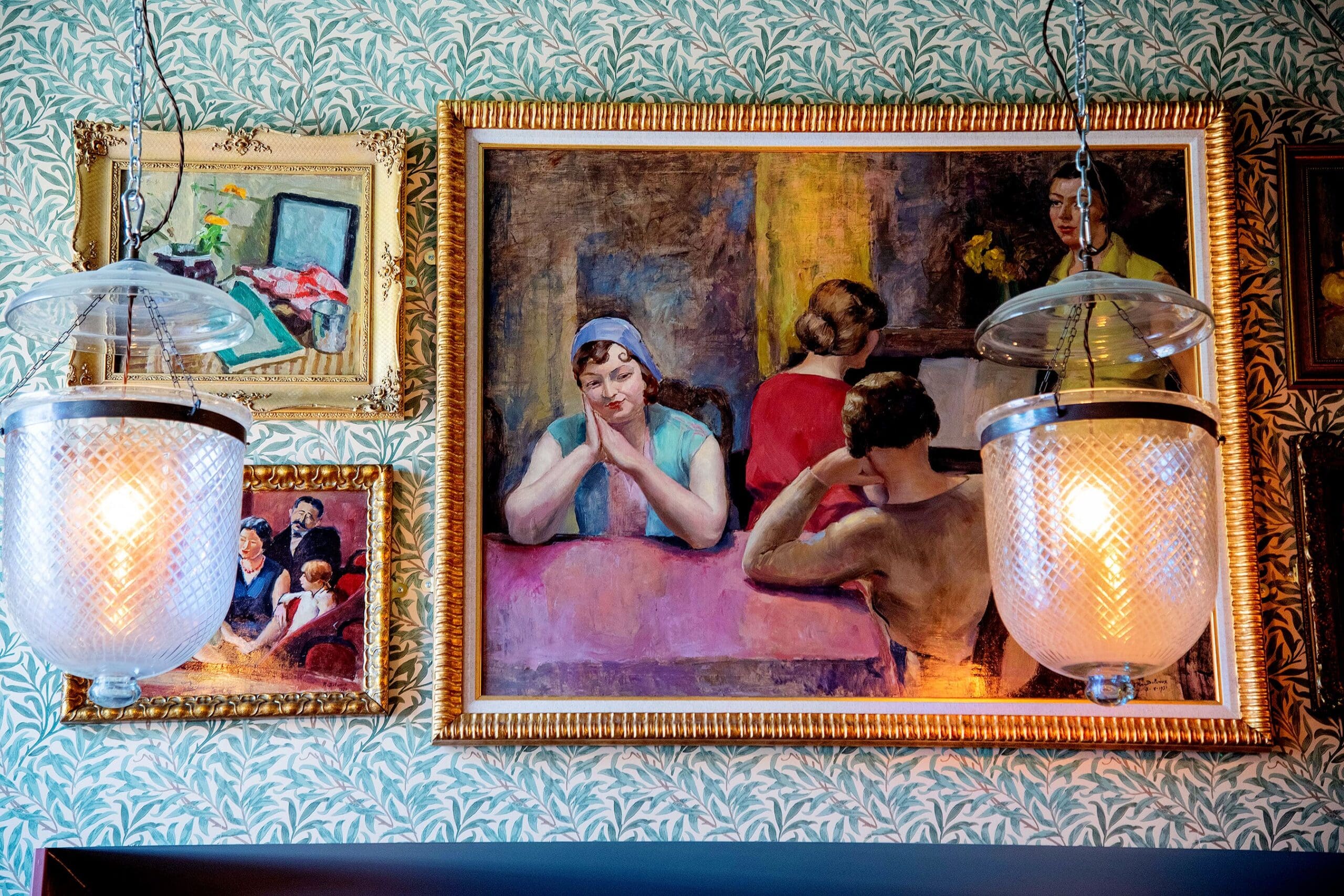 Two ovino glass pendant lights hang in front of a wallpapered wall adorned with framed paintings, including a central artwork of people at a pink table, one woman with folded hands appearing thoughtful.