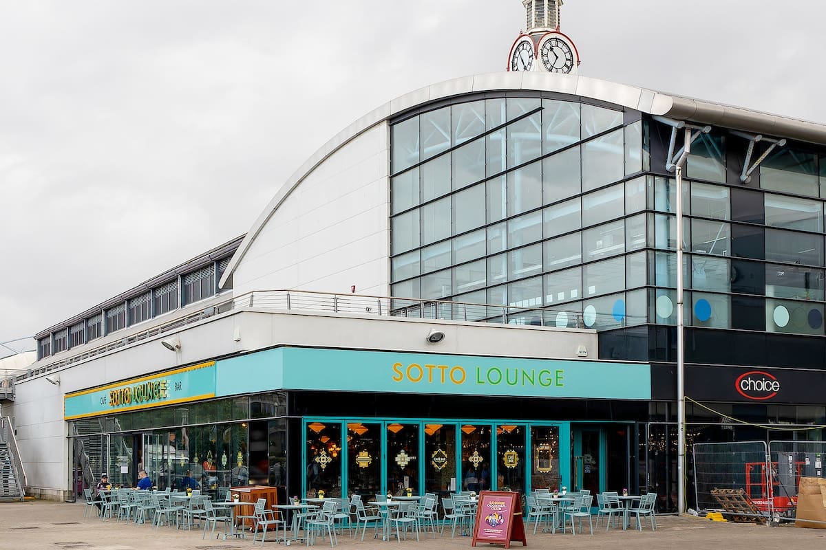 A modern, glass-fronted building houses Sotto Lounge, with vibrant turquoise outdoor seating and bold yellow signage. The Sotto clock tower stands on the roof, set against an overcast sky.