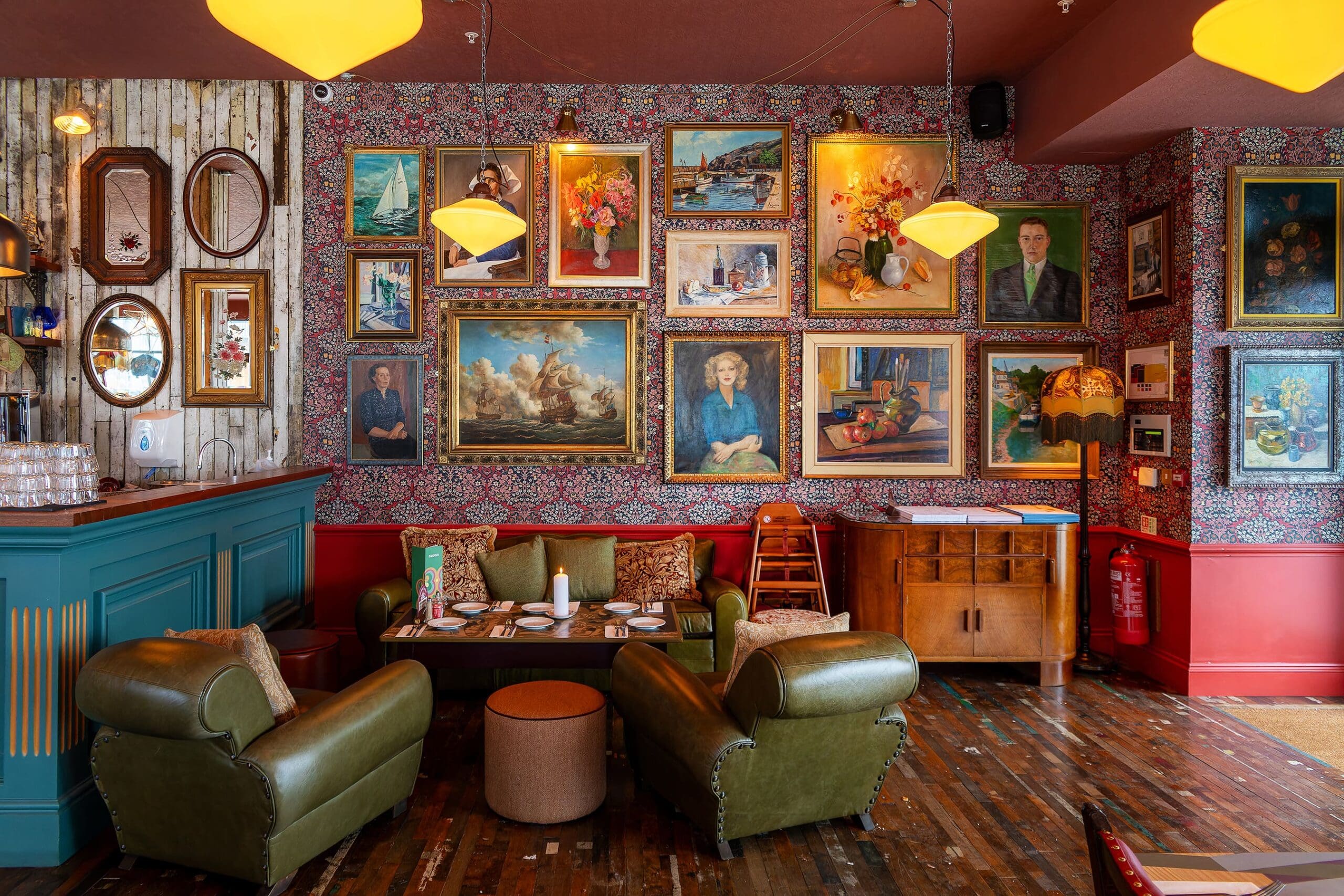 A cozy vintage-style lounge by Sotto with green leather sofas, a wooden floor, and colorful patterned wallpaper covered in framed paintings and portraits. Warm lighting hangs from the ceiling above.