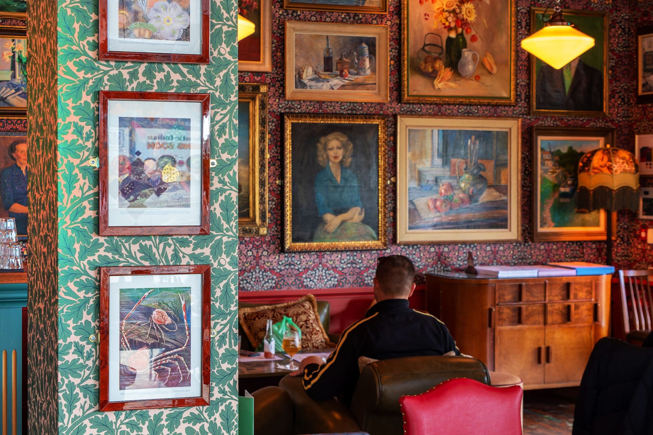 A cozy cafe interior with Sotto-patterned wallpaper, many framed paintings on the walls, and a person sitting on a chair facing away. Warm lighting creates an inviting atmosphere, and tables are set for guests.
