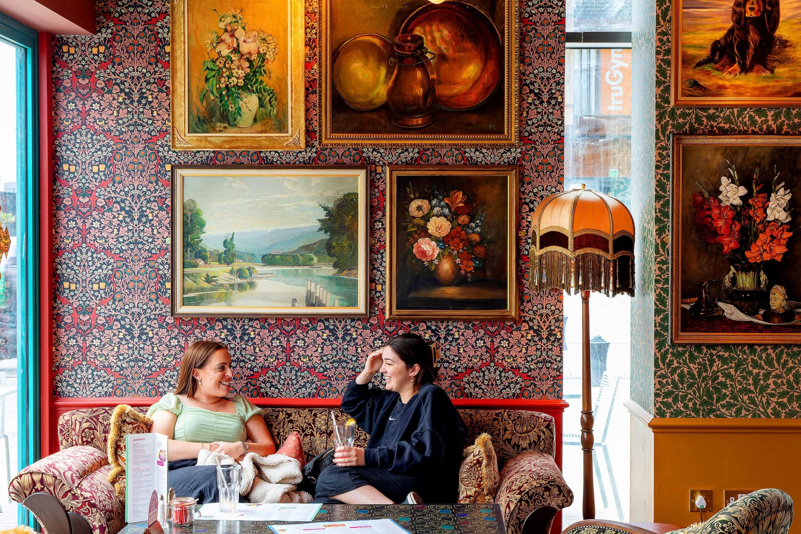 Two women sit sotto on a vintage sofa in a cozy, art-filled room with patterned wallpaper, ornate lamps, and framed paintings of landscapes, flowers, and animals, quietly engaged in conversation and enjoying drinks.
