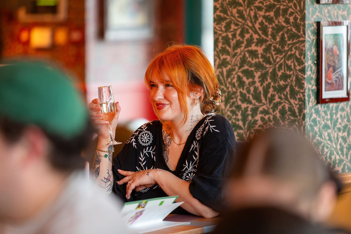 A woman with orange hair and tattoos is smiling and holding a glass of Soffio, sitting at a table in a lively, patterned restaurant. Other people and wall art are visible in the background.