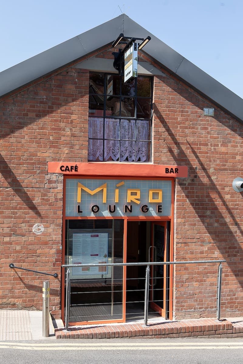 The entrance of Miro Lounge, a café bar in a brick building with large glass doors, orange Miro signage, and a metal handrail leading to the door. A menu is displayed by the entrance.