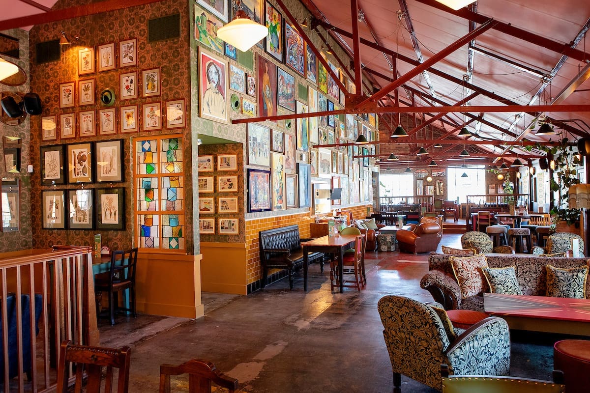 A vibrant, eclectic cafe interior with colorful patterned armchairs, wooden tables, and walls covered in Miro-inspired art and portraits. The space has high ceilings with exposed beams and large windows letting in natural light.