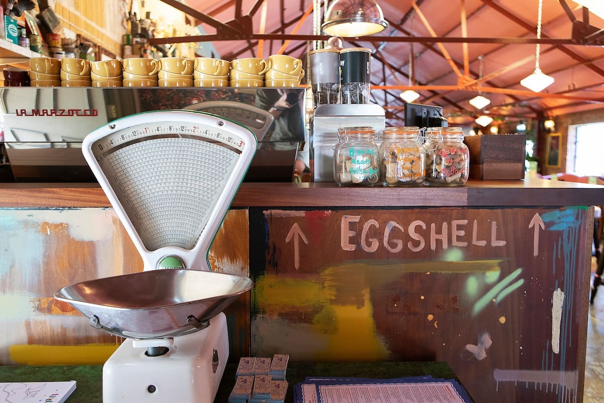 A vintage kitchen scale sits on a colorful Miro-inspired counter in a café. Behind it are stacked cups, jars with labels, a coffee machine, and a sign reading "EGGSHELL" with arrows pointing up. Warm lighting fills the space.