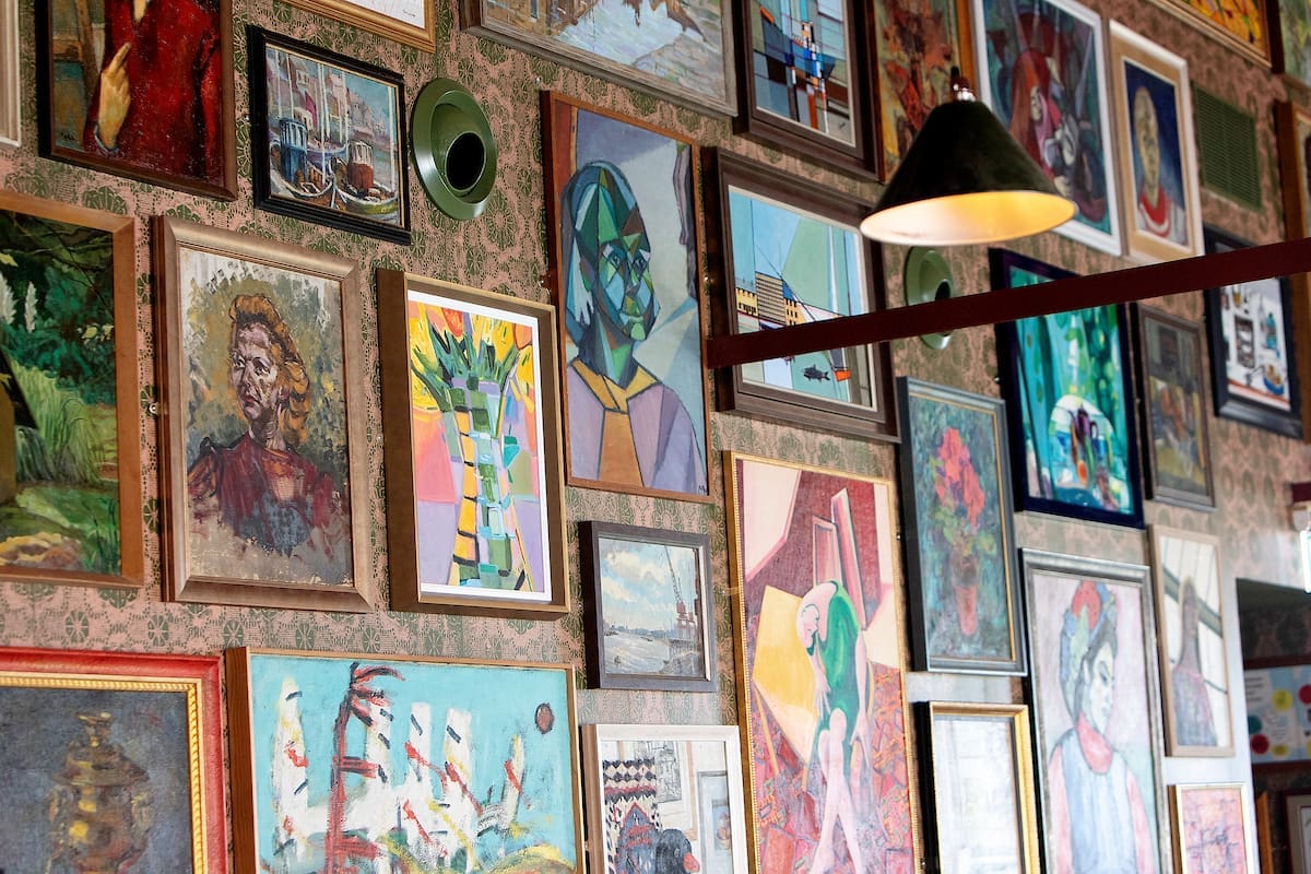 A wall filled with various colorful framed paintings, including portraits, abstract art inspired by Miro, landscapes, and still lifes, under a hanging lamp in a cozy, art-filled room.