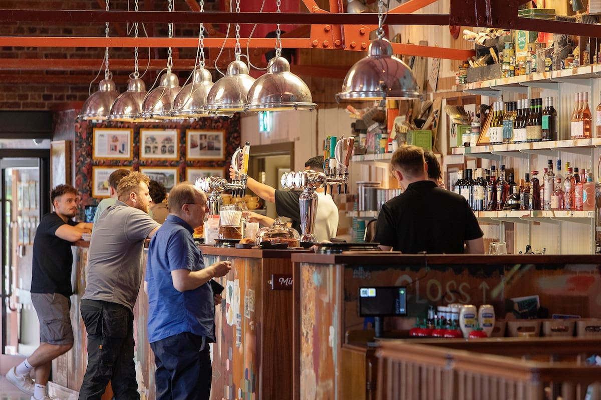 Several people stand and sit at a bar counter inside a cozy, well-lit pub with hanging lights, exposed beams, and shelves filled with bottles and glassware. A bartender serves drinks behind the counter, while a group sketches ideas on Miro at one end.