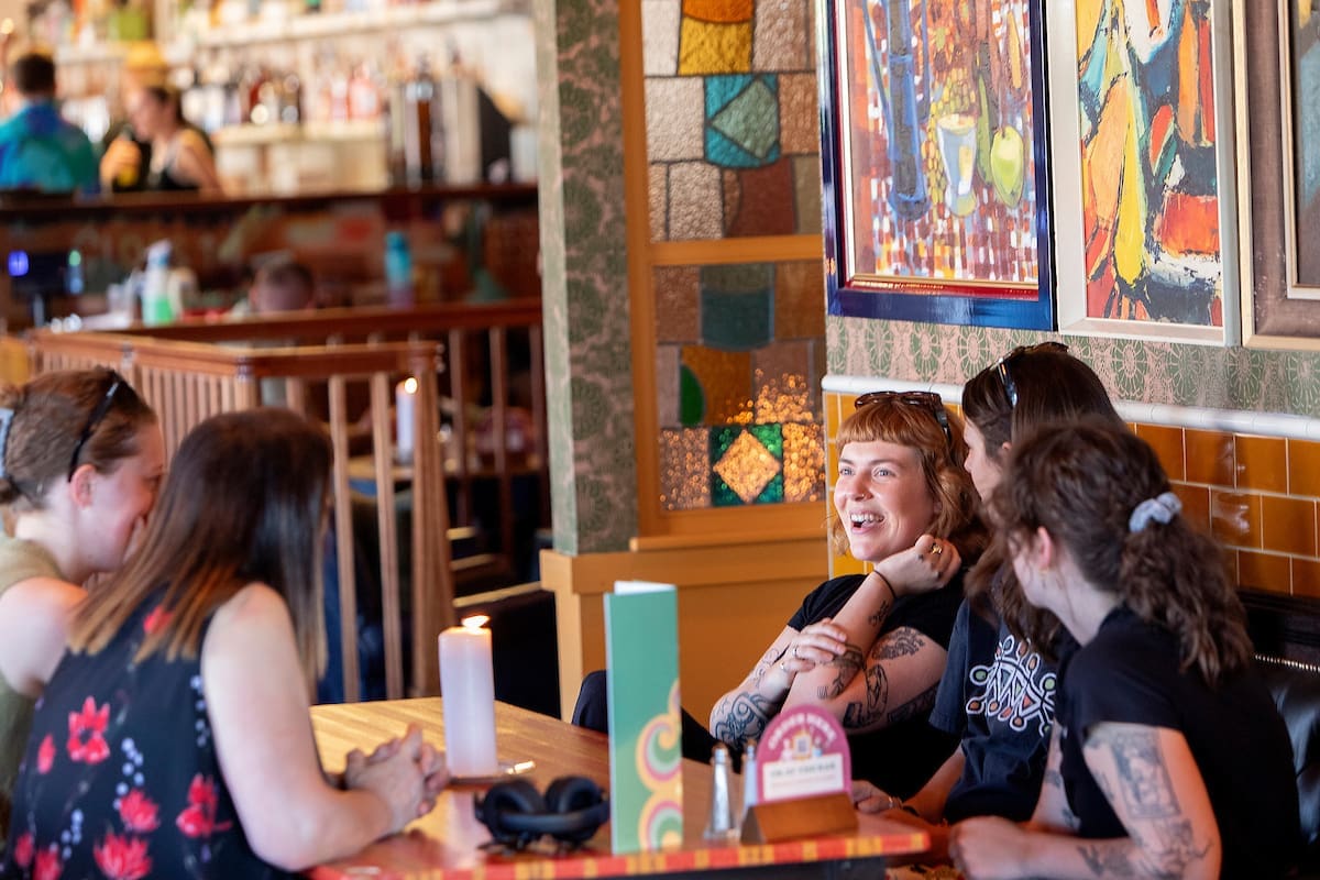 Four women sit and chat at a restaurant table, smiling and laughing. Colorful Miro-inspired artwork and stained glass decorate the walls, creating a lively and warm atmosphere.