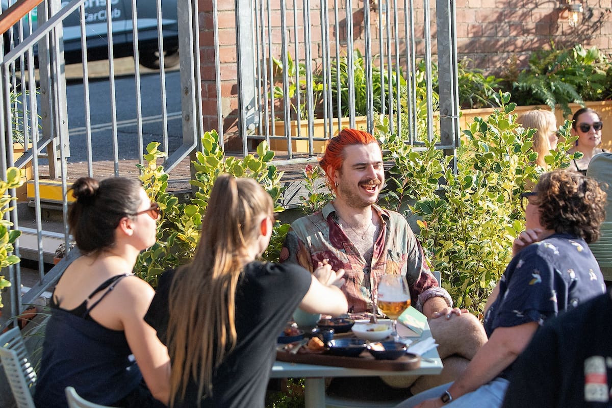 Four people sit around an outdoor table at a café or bar, talking and laughing. One person with bright orange hair is smiling widely. Plates and drinks are on the table, and sunlight brightens the lively, Miro-inspired scene.