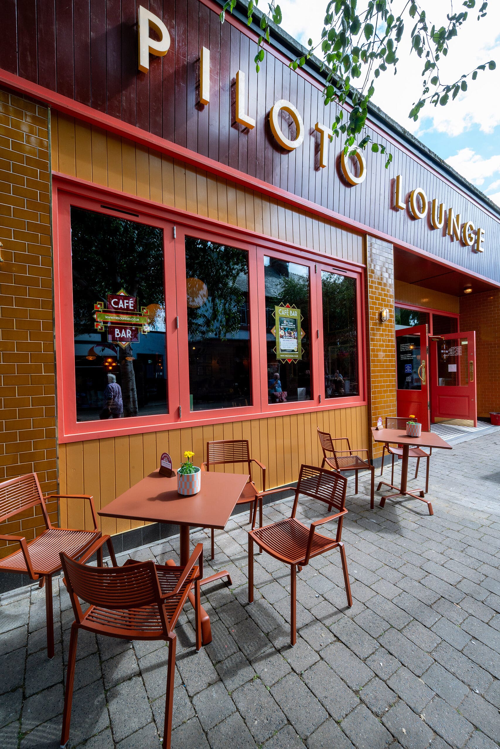 Outdoor seating area with brown tables and chairs in front of Piloto Lounge, a vibrant café and bar with red-framed windows. The yellow and red Piloto facade stands out amidst tree branches and cobblestone pavement.