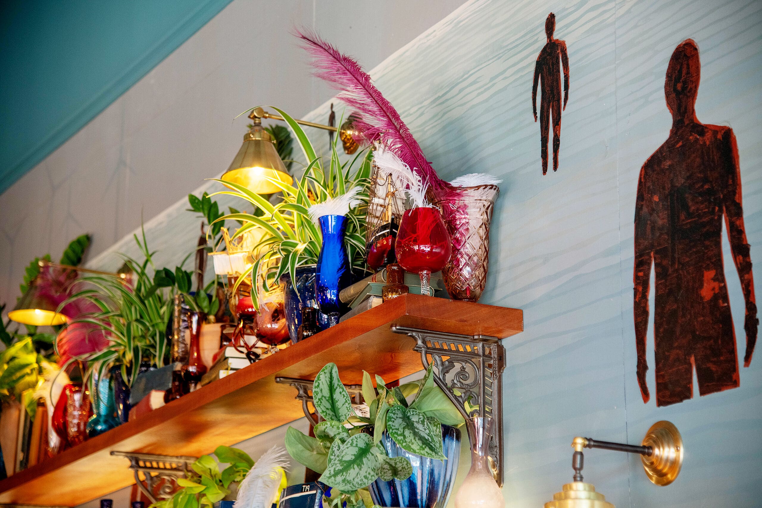 A wooden Piloto shelf with assorted potted plants, colorful glass vases, and pink feathers, mounted on a wall with painted silhouettes of human figures in the background. Warm lighting illuminates the display.
