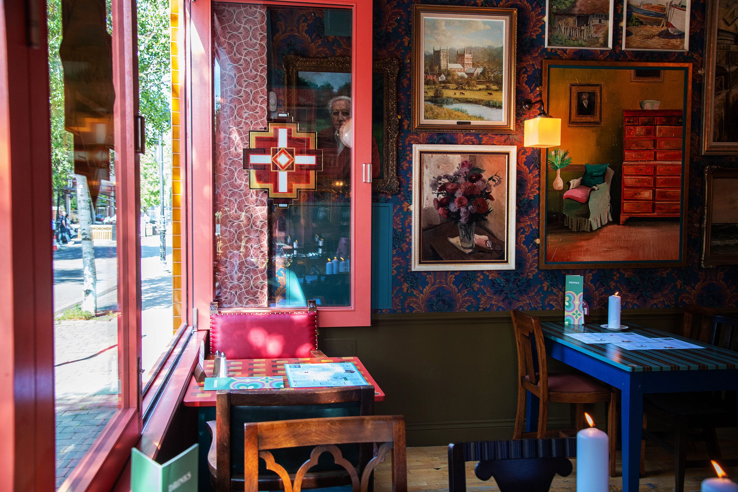 A cozy café corner, Piloto-inspired, with colorful walls adorned with framed paintings, wooden tables and chairs, a red cushioned seat by the window, and sunlight streaming in from outside.