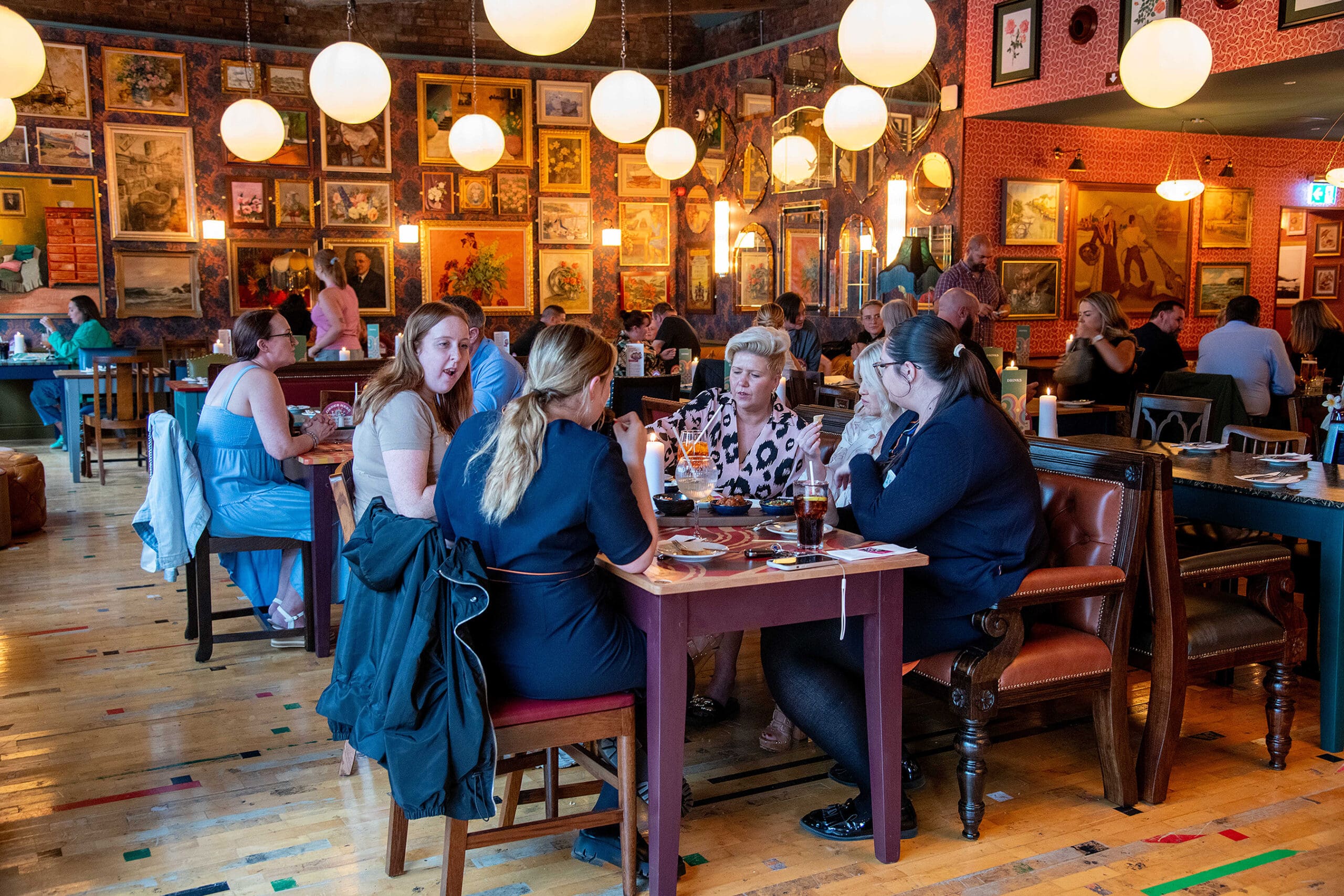 People dine and talk in Piloto, a lively restaurant with colorful paintings on the walls and globe pendant lights hanging from the ceiling. Several groups sit at wooden tables, enjoying food and drinks in a cozy, artistic atmosphere.