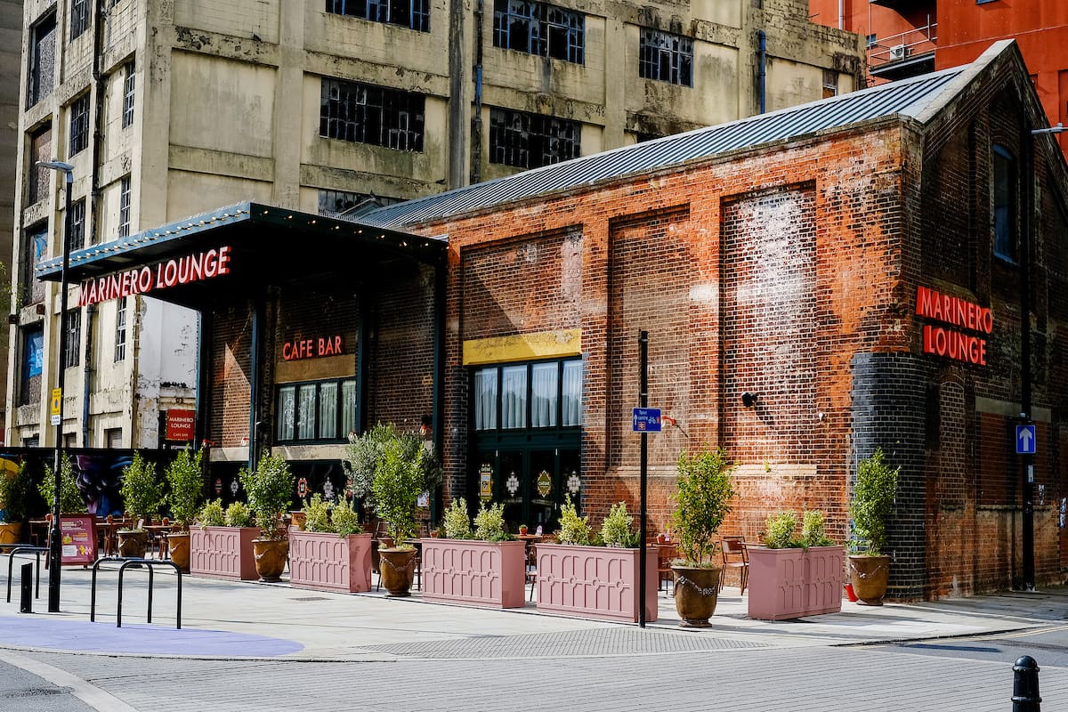 A brick building with a metal roof houses the Marinero Lounge café bar, featuring outdoor seating, potted plants, and bright red signage. The urban marinero setting is surrounded by tall, older buildings nearby.