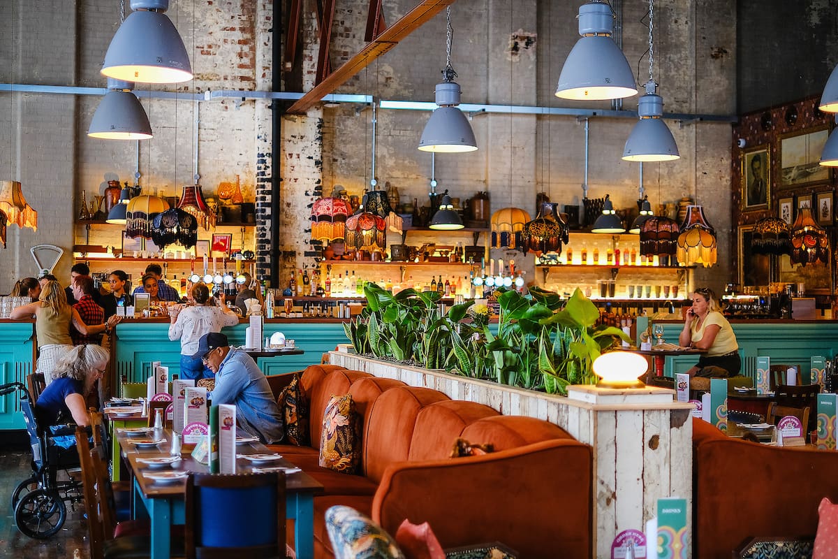 A vibrant café with eclectic decor, orange couches, green plants, and vintage lamps. The cozy, lively atmosphere features exposed brick walls and pendant lights—a perfect spot for a marinero to relax among friends or enjoy a break at the counter.