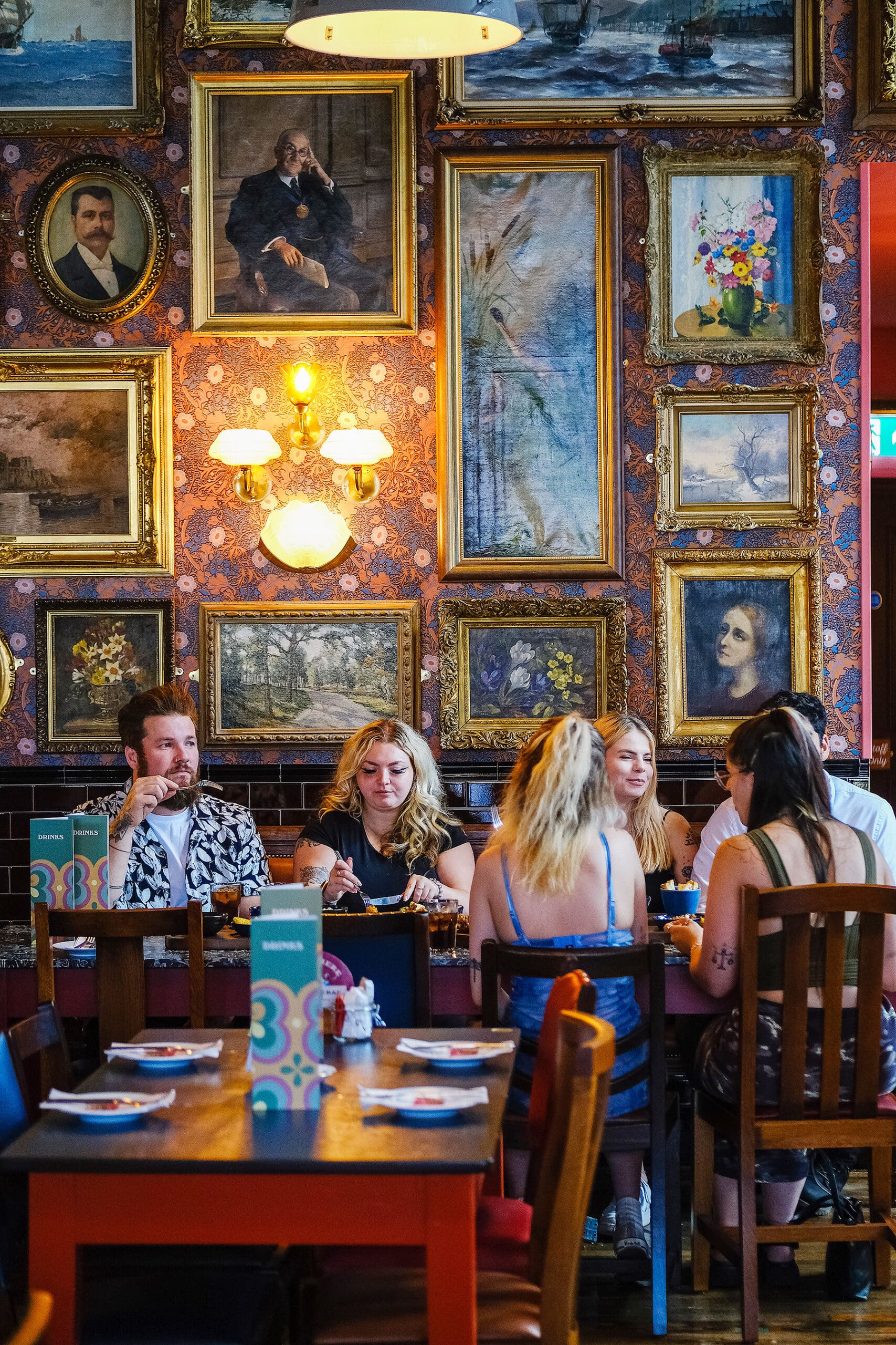 A group of people sit around a table in a colorful, marinero-themed restaurant decorated with ornate framed paintings and portraits on patterned wallpapered walls. They are engaged in conversation and enjoying their meal.