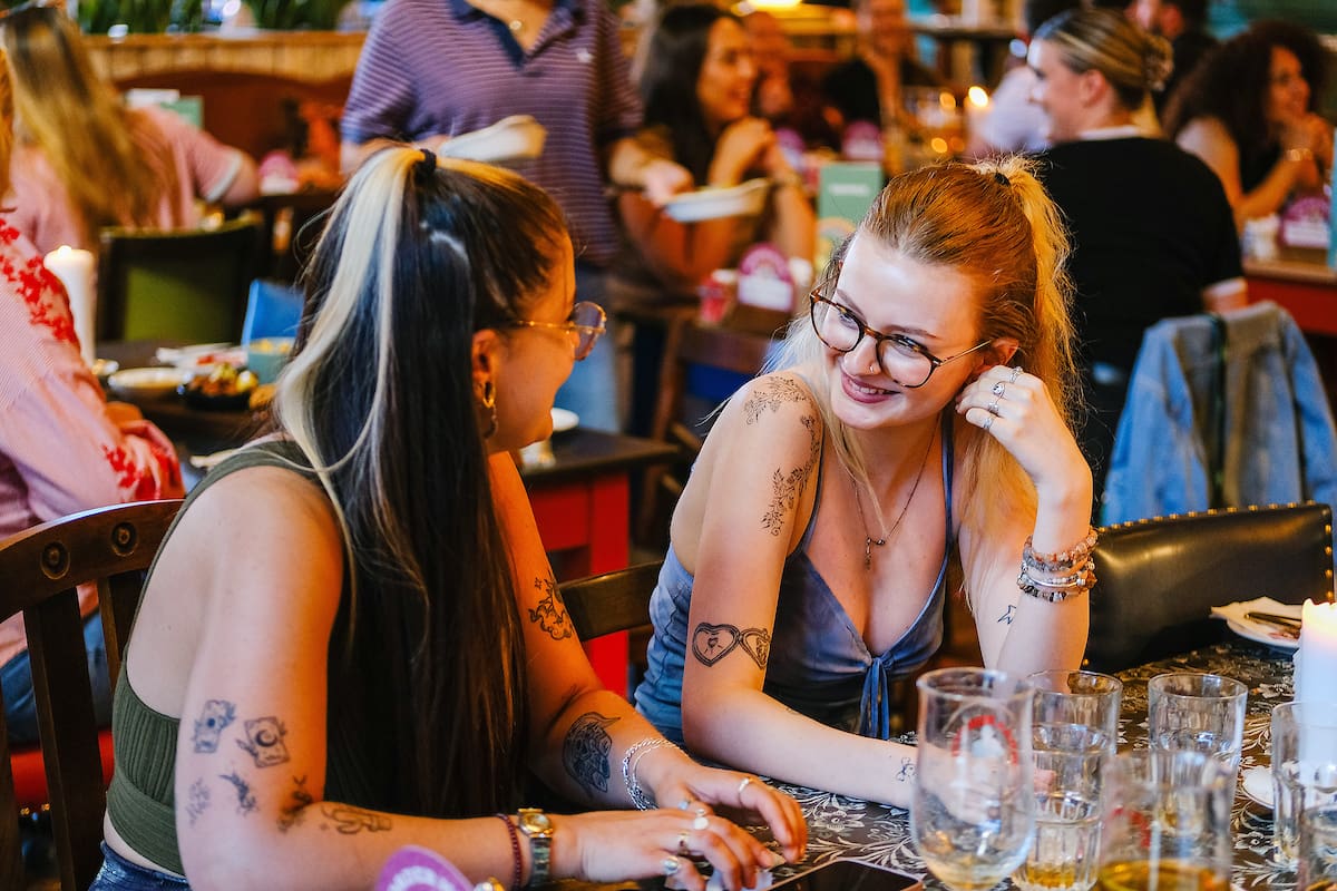 Two women with tattoos and glasses sit at a restaurant table, smiling and talking. Glasses and dishes are on the table, while a lively marinero-themed décor fills the colorful background with energy.