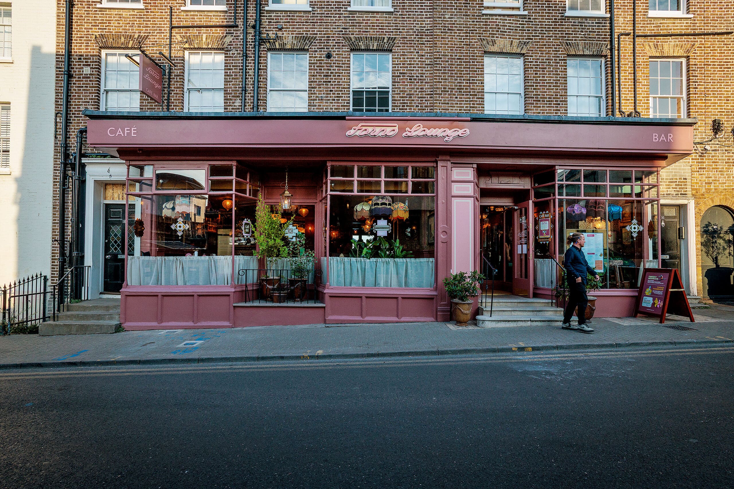 A person walks past a pink-painted cafe, bar, and forno with large windows, plants, and colorful hanging decorations inside. The brick building stands out on an empty street.